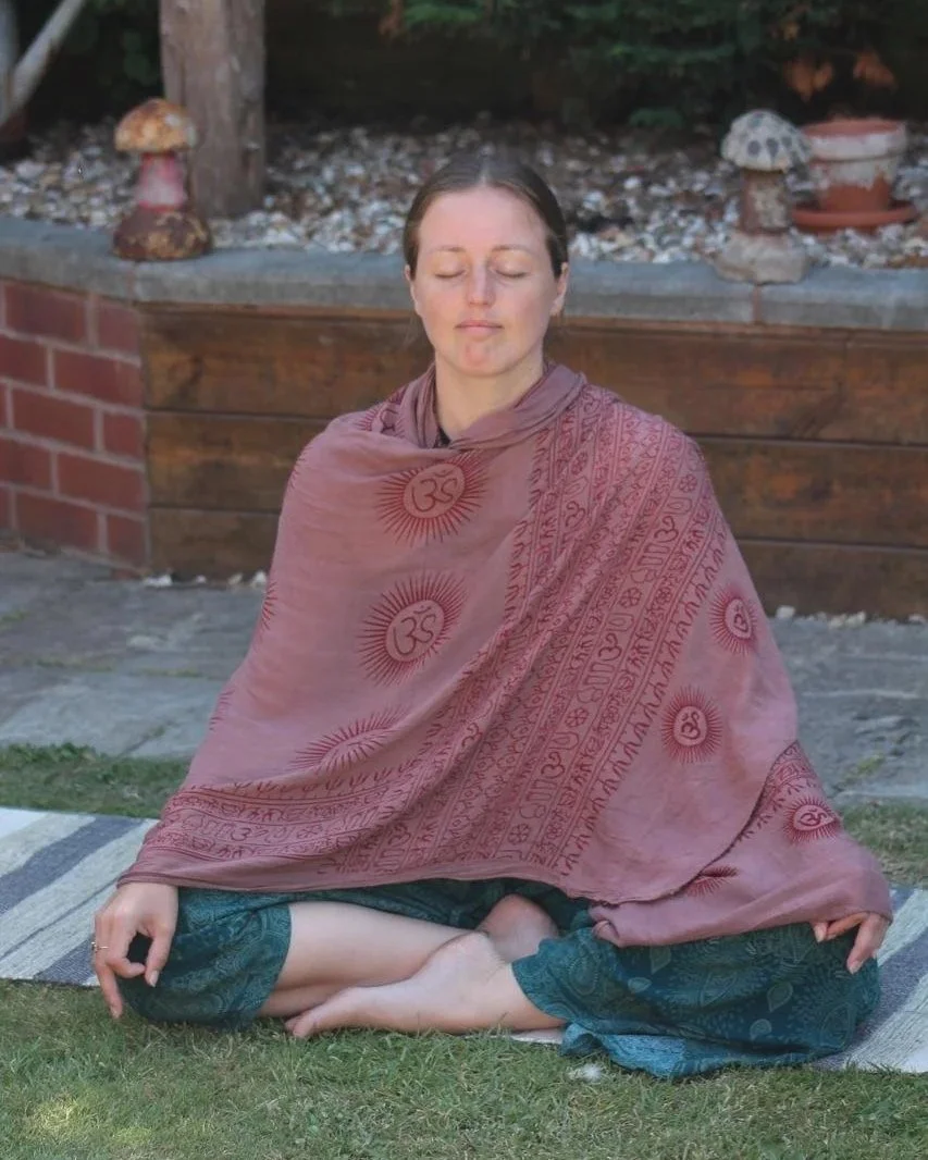 A woman sitting cross-legged on a yoga mat, doing 1:1 and group pranayama which is breathing with retention from India. For the yoga she is wearing a black sports bra and gray shorts, with her eyes closed and hands in a mudra.