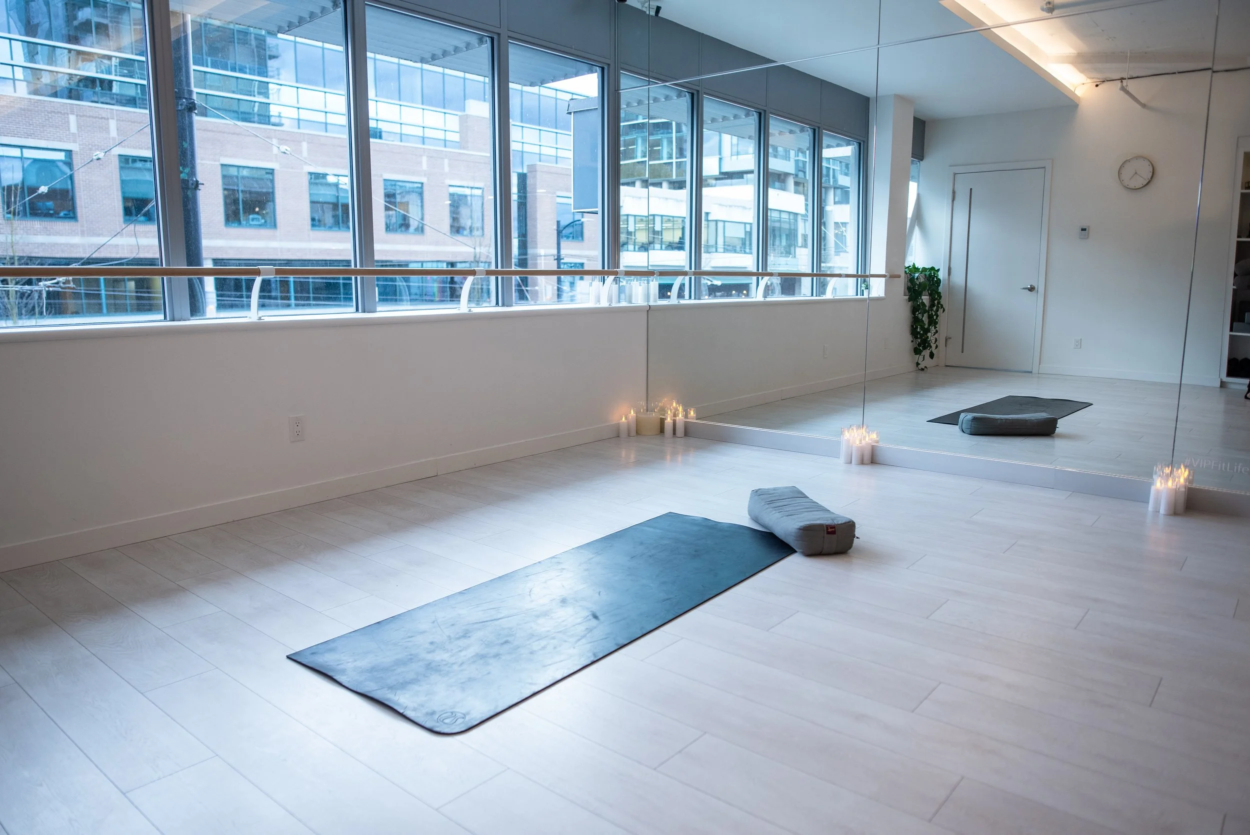 A spacious yoga or meditation studio with large windows for 1:1 private yoga and group sessions, light wooden floors, yoga mats, rolled towels or blankets, candles, a mirror, and a clock on the wall.