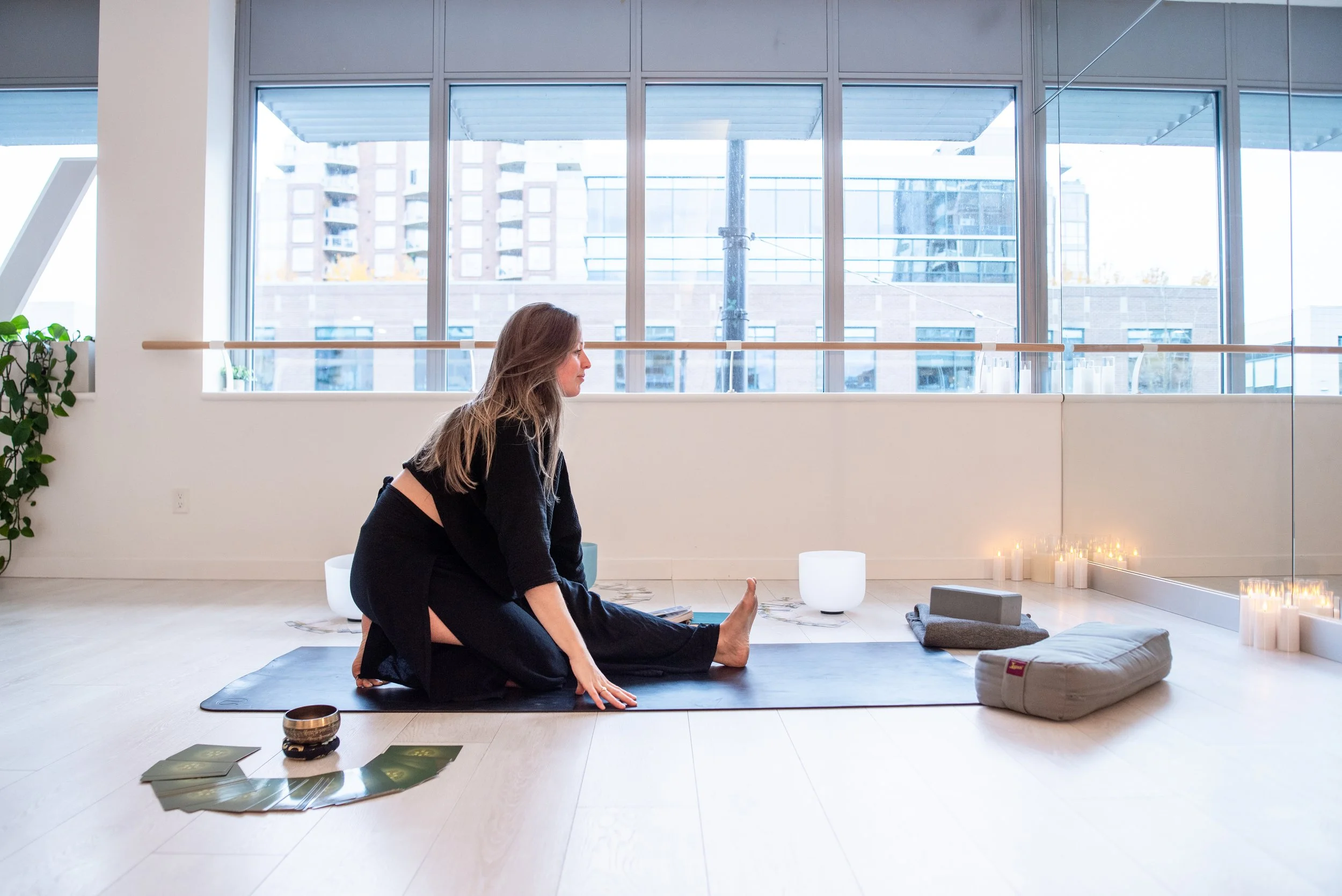 A woman practicing 1:1 and group yoga on a black mat inside a bright room with large windows overlooking city buildings. She is in a seated forward bend pose in half monkey and reaching for her feet, with candles and yoga props around her.