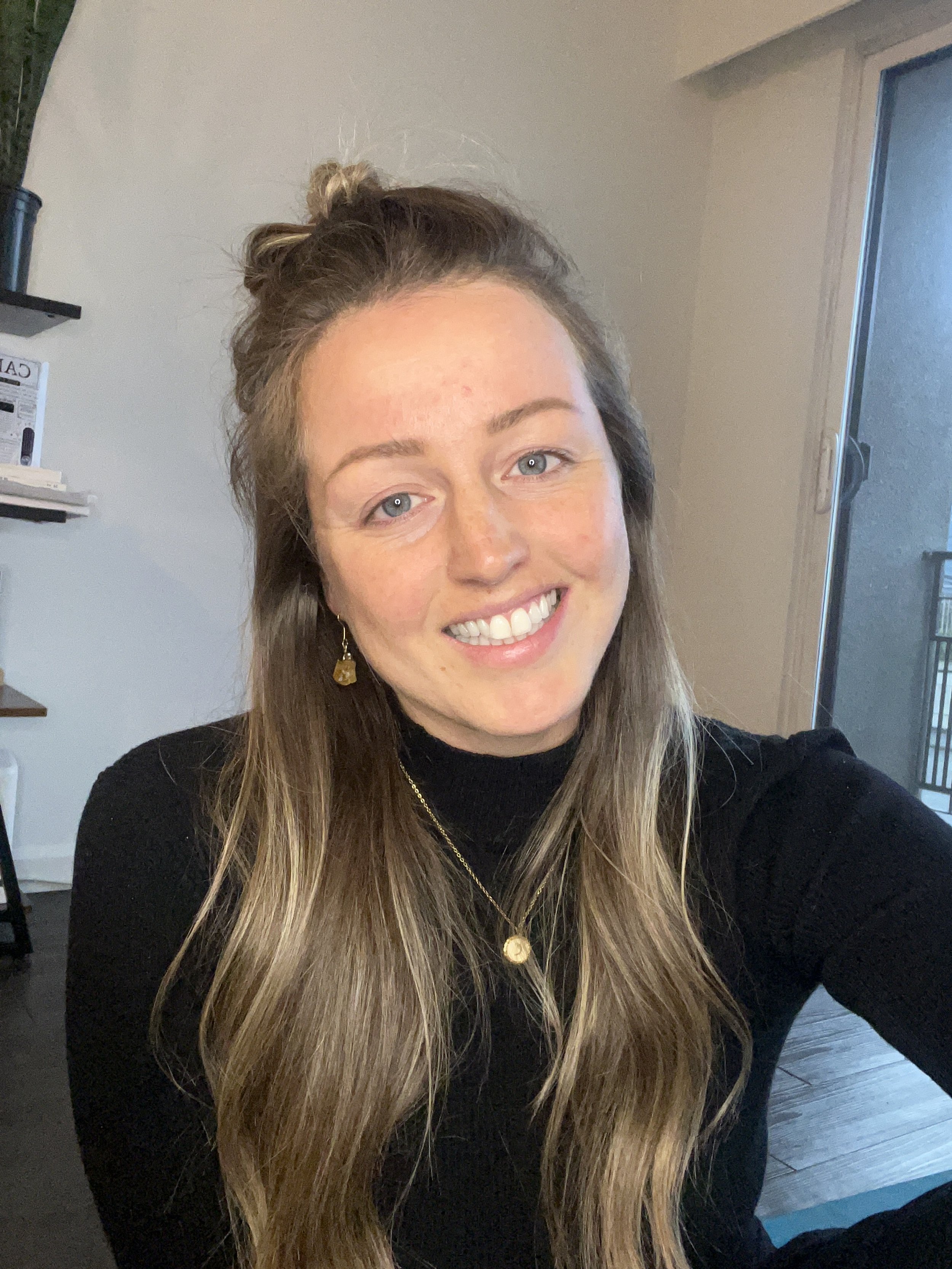 A woman with long, wavy hair taking a selfie indoors, smiling, wearing a black top and gold jewelry, with a sliding glass door and some shelves in the background.