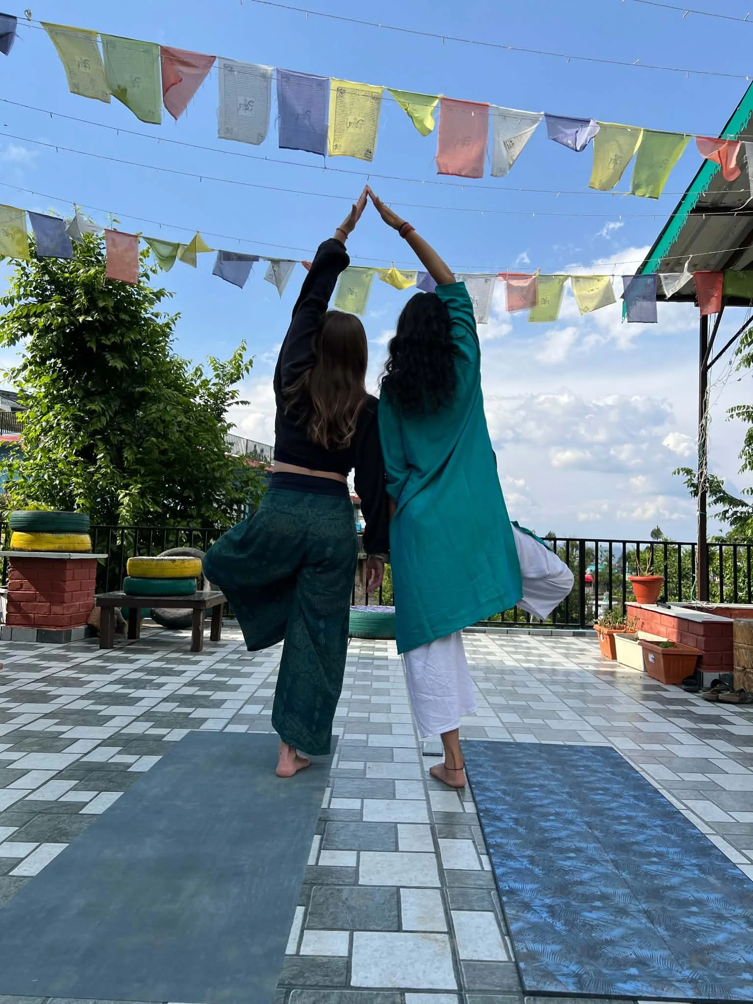 A Eastern Indian woman and a Western English woman practicing yoga on a rooftop terrace together with prayer flags overhead, trees and a blue sky in the background in India.