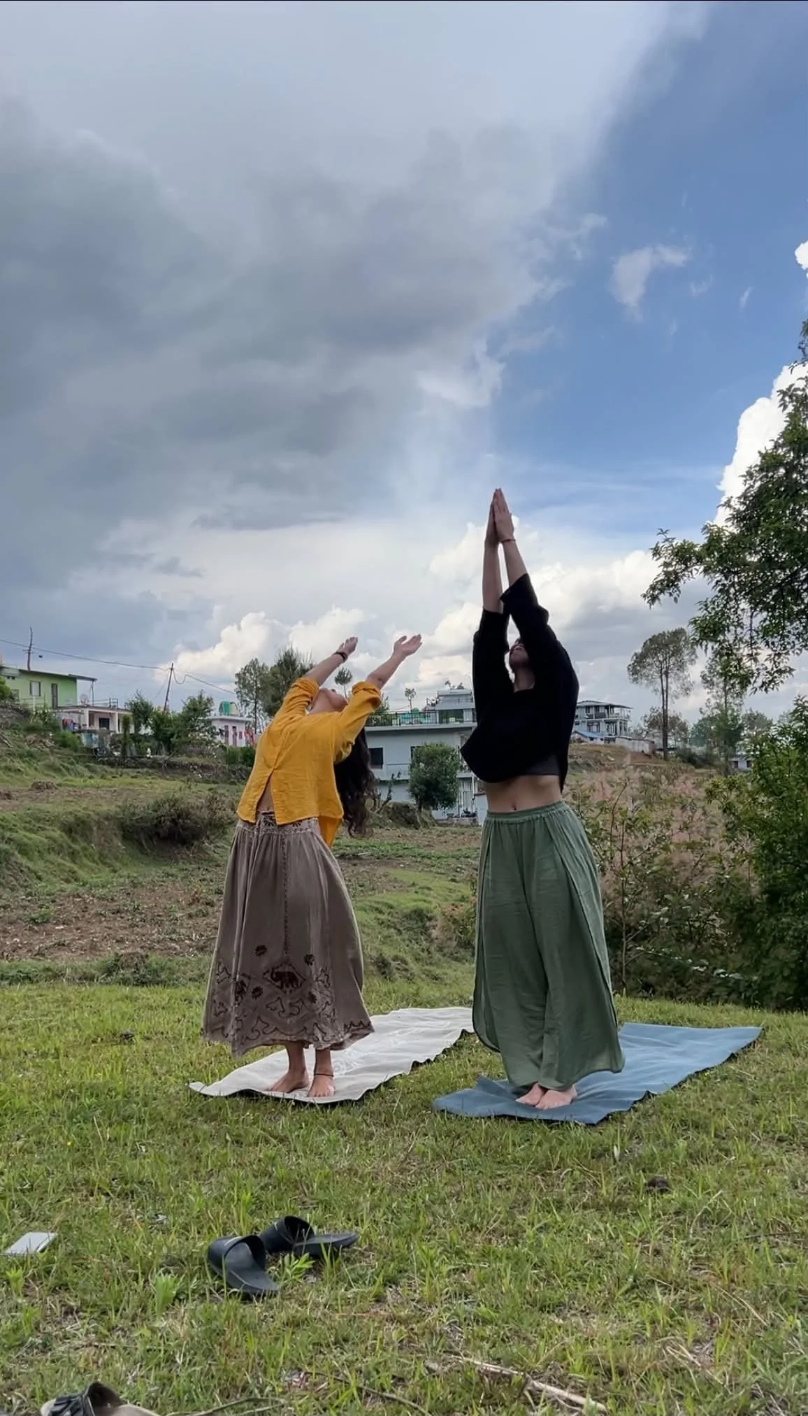 A Eastern woman and Western woman practicing yoga outdoors on yoga mats in a grassy field, with one raising their hands above their head and the other reaching upward with arms extended, showing the indian culture and traditions.