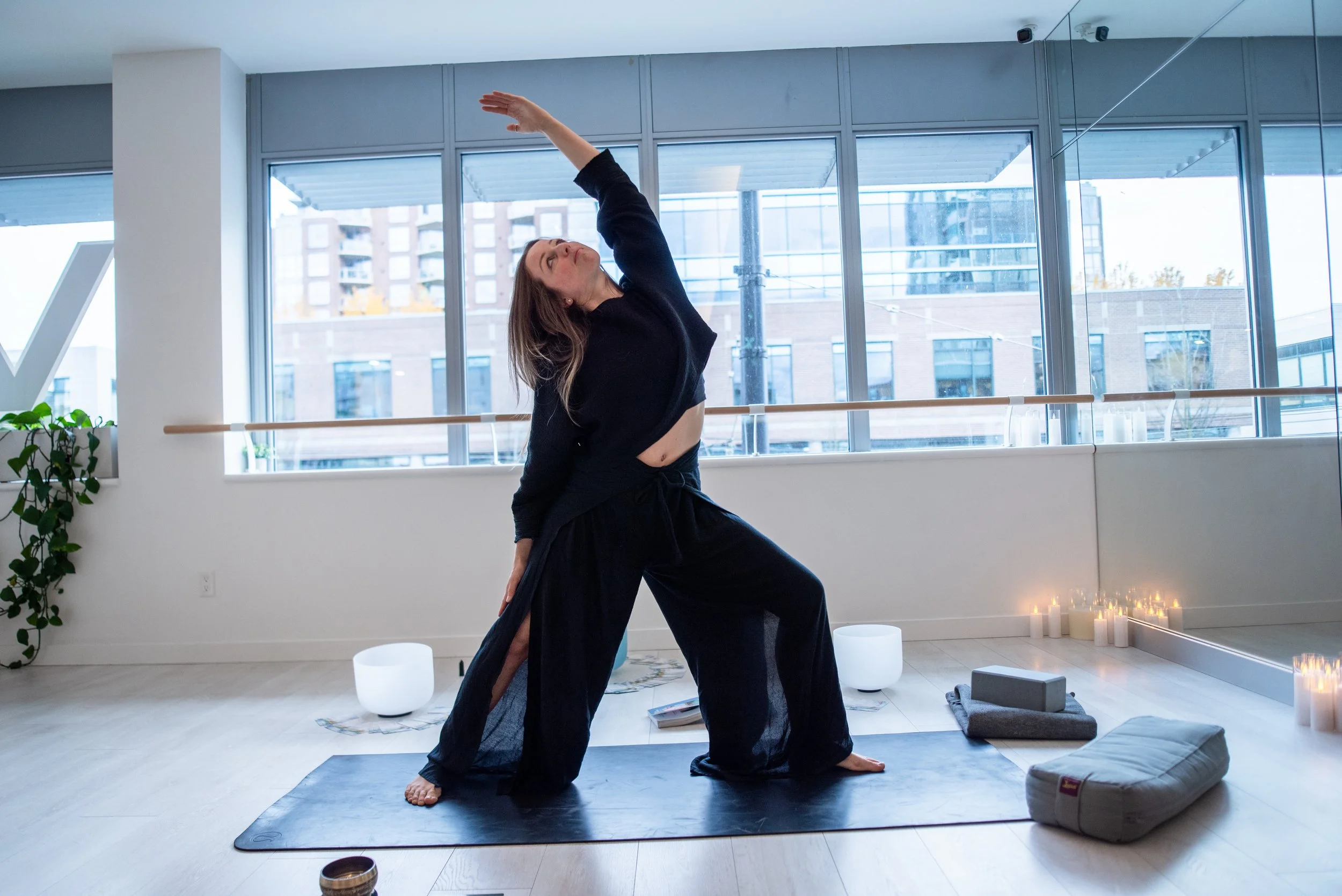 A woman practicing yoga or stretching in a bright, modern studio with large windows and cityscape view. She is on a black yoga mat, wearing black clothing, with candles and meditation items around.