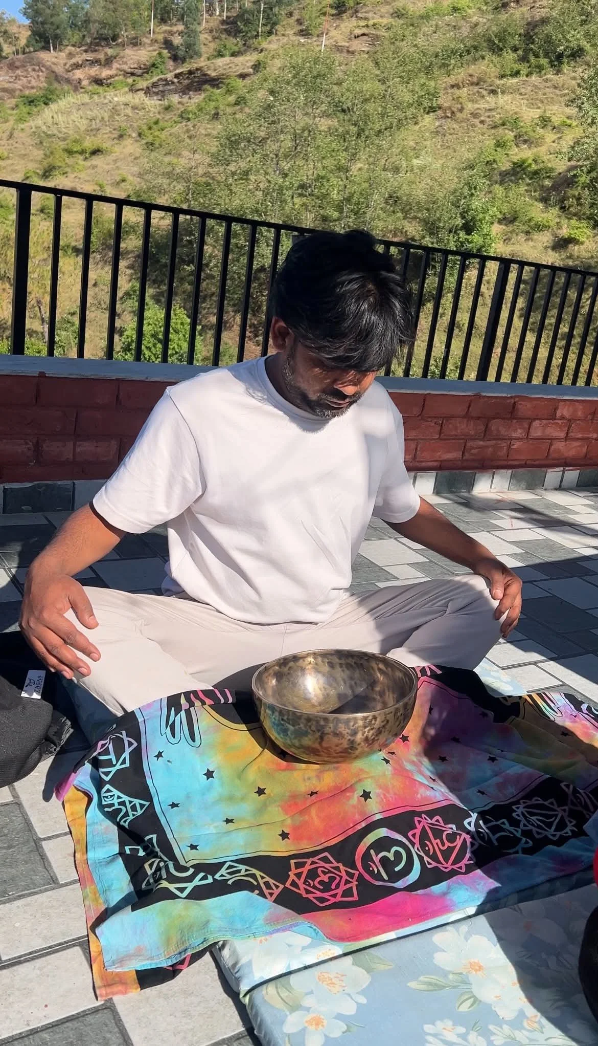 A man doing 1:1 sound healing with tibetan bowls to promote healing from a injury. He has a colourful buddha throw and he is wearing a white T-shirt and beige pants, with trees and a hillside in India.