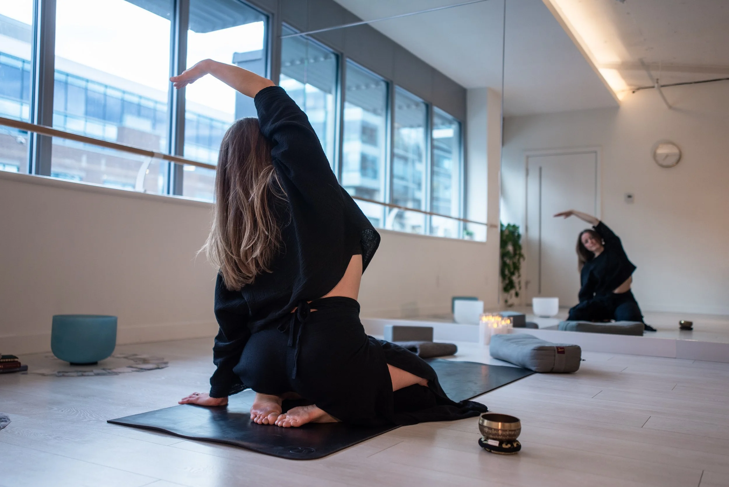 A women practicing  1:1 and group yoga in a bright studio near large windows, one in the foreground on her knees, the other in the background stretching with one arm raised.
