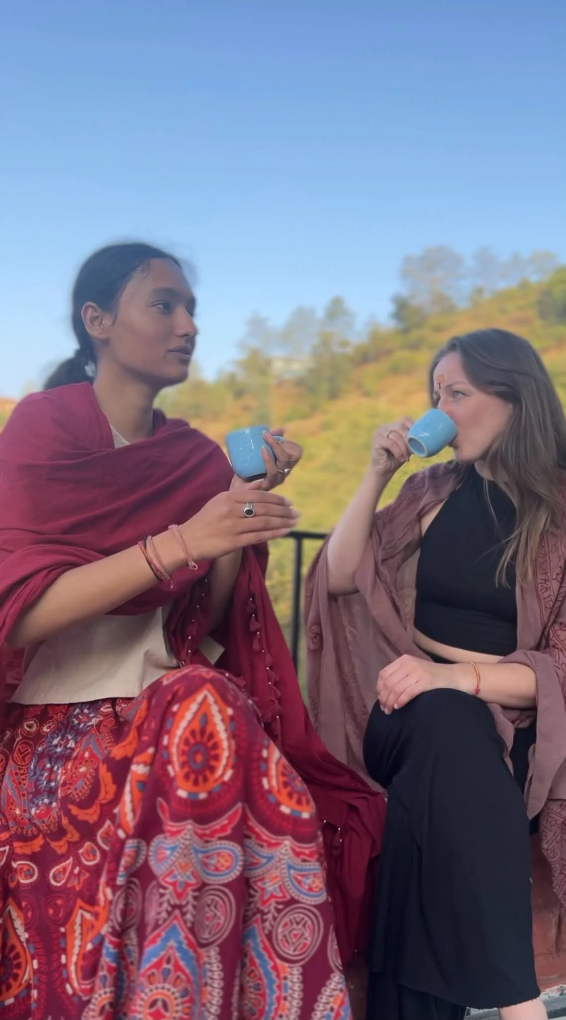 A Eastern women and Western woman sitting outdoors, one with dark hair and the other with light brown hair, drinking tea from blue cups discussing cultural exchange and indian tradtions and philosophy against a background of trees and a blue sky.
