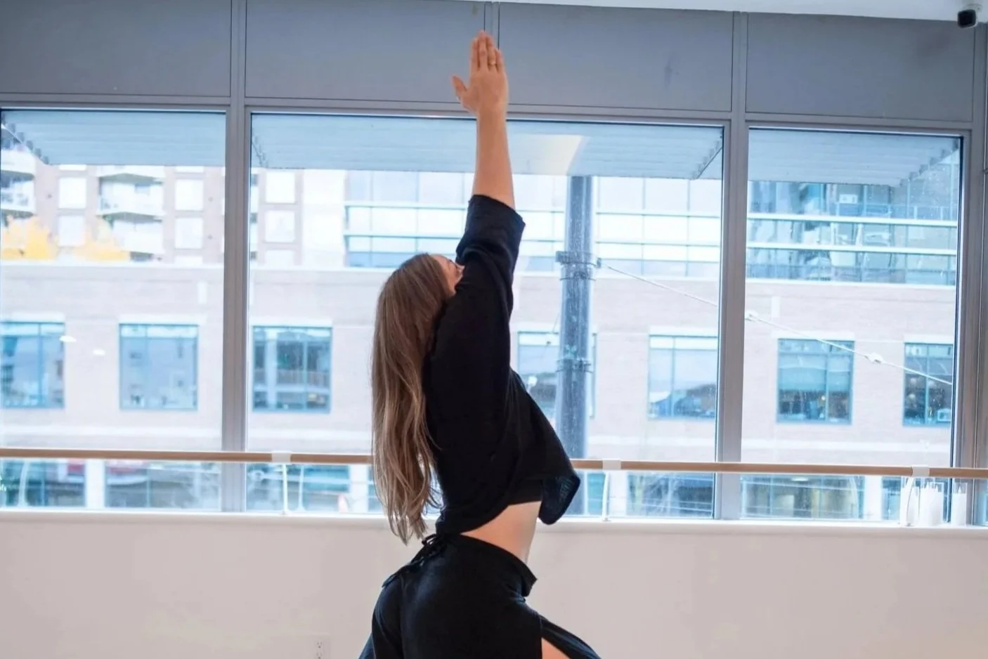 A young woman practicing yoga in a studio doing warrior 1, also known as Virabhadrasana 1 position, wearing black clothing, with her arms raised upward and head tilted back.