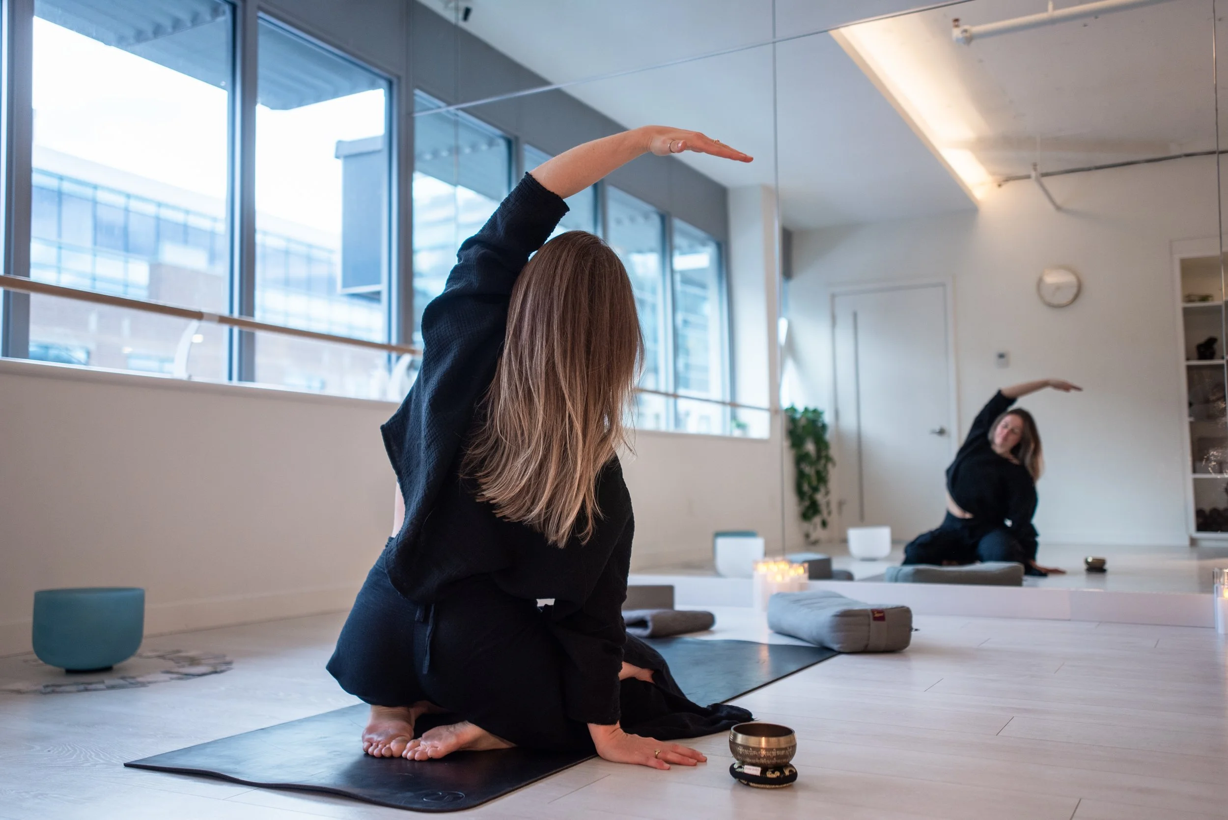 A woman practicing yoga 1:1 and group sessions in a modern studio with large windows, one woman kneeling with her arm raised and the other doing a split pose in the background, stretching.
