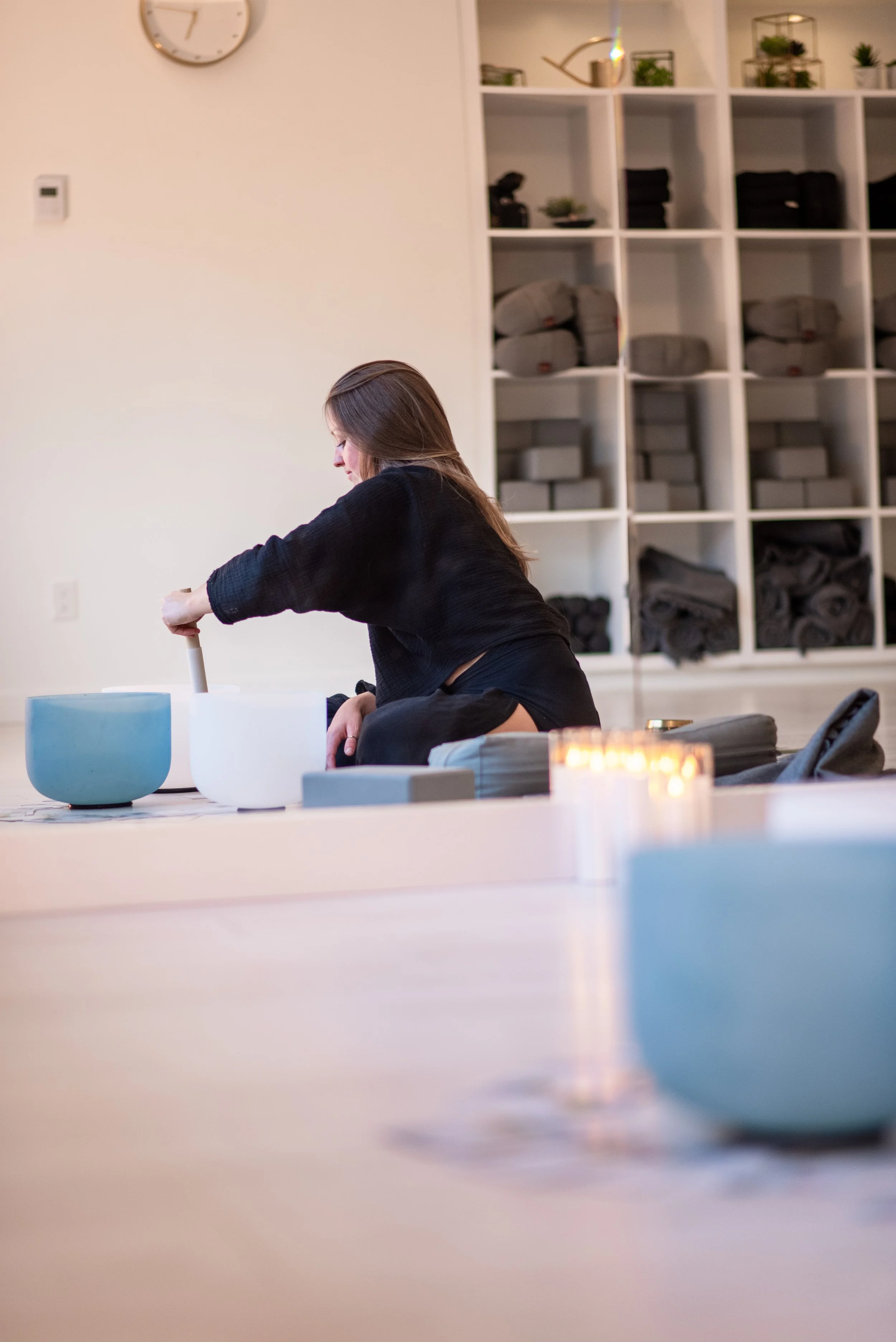 A woman with long brown hair doing 1:1 private and group sound healing with relaxtion crystal sound bowls. inside a room with white walls and shelves filled with neatly folded dark and gray towels and some small potted plants.