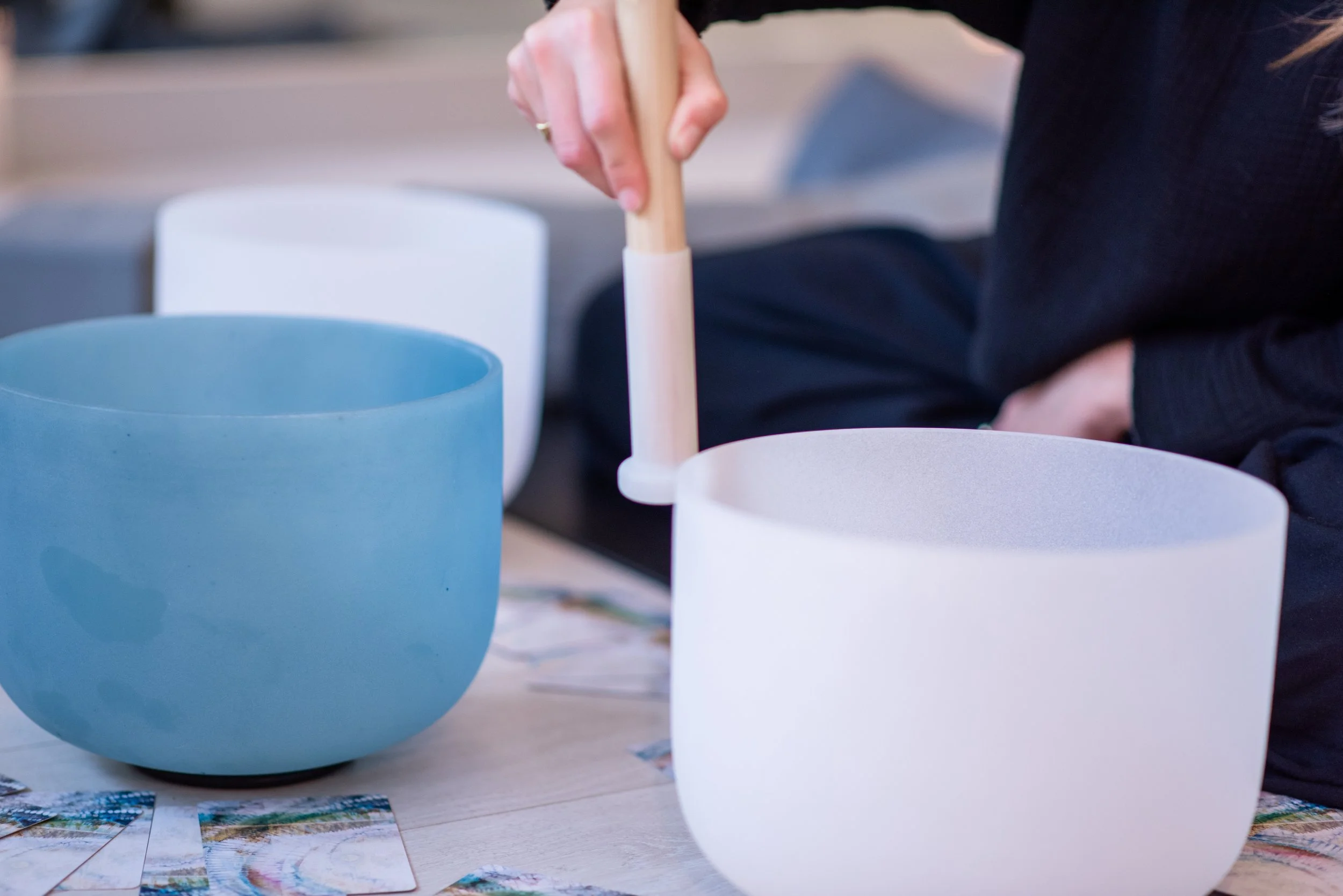 Person doing 1:1 and group sound healing, playing crystal sound bowls with a mallet or hammer near large, rounded, crystal bowls in various colors including blue and white, with colorful photographs spread on the table nearby.