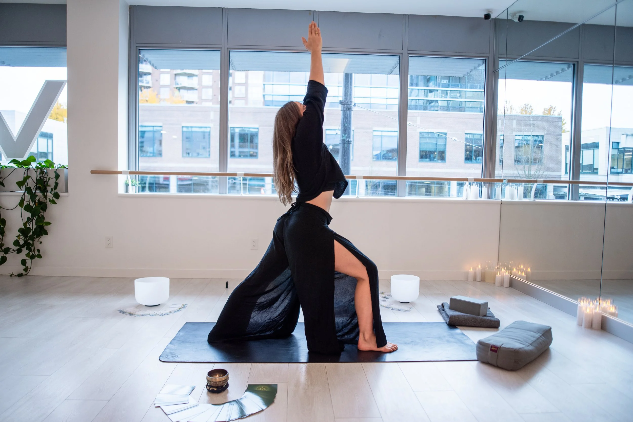 A women practicing 1:1 and group yoga indoors, in warrior 1 position with her arms ovr head stretching, a classic yoga position. The room has large windows, dirndl lighting, and candles on the floor.