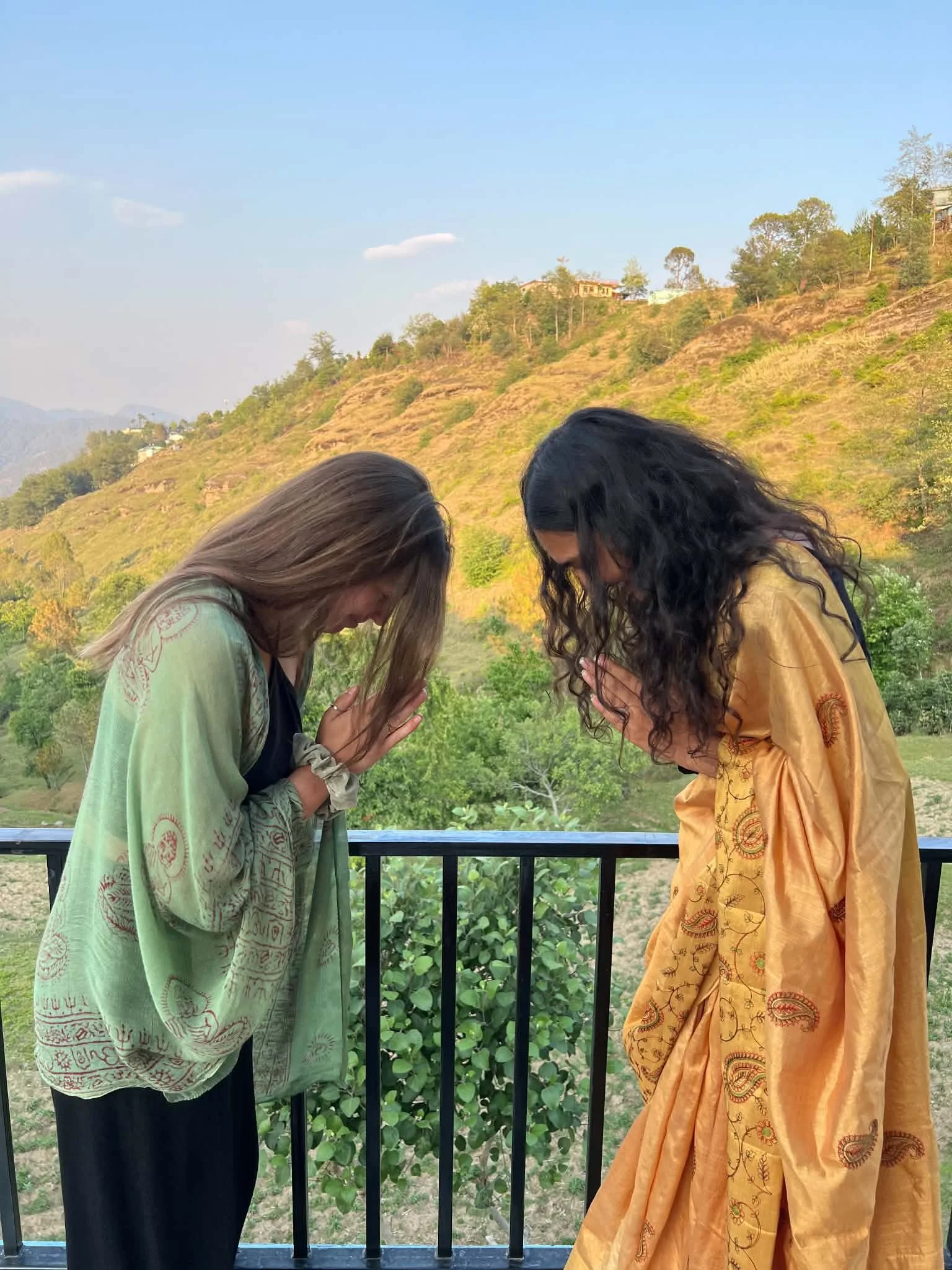A Western women  and Eastern woman bowing with their heads lowered and eyes closed in Anjali Mudra, standing outdoors on a balcony with a scenic backdrop of hills, trees, and a partly cloudy sky in India.