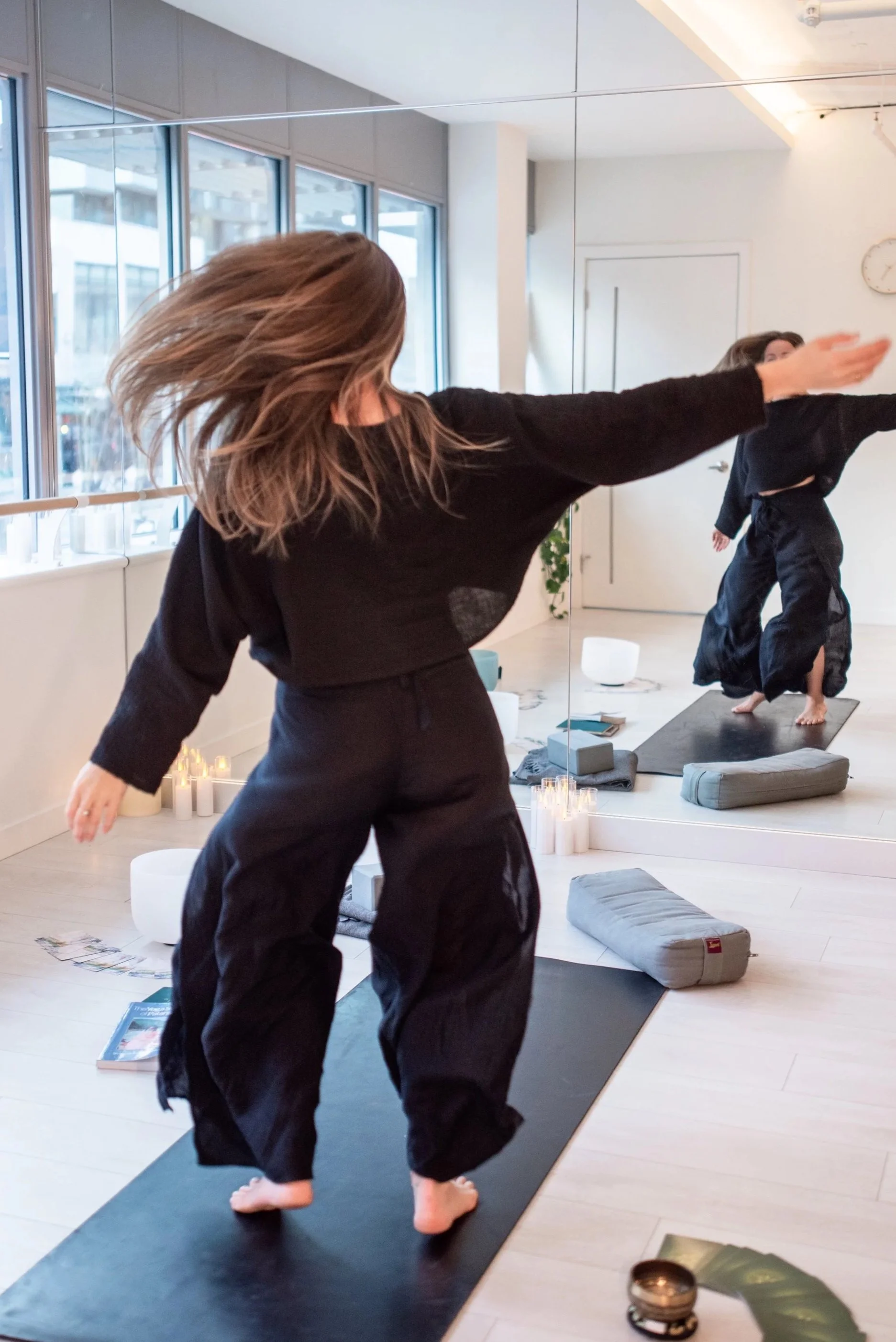 Person practicing 1:1 online and in-person yoga and intutie movement in front of a mirror in a bright, minimalistic studio with large windows, candles, and yoga mats on the floor.