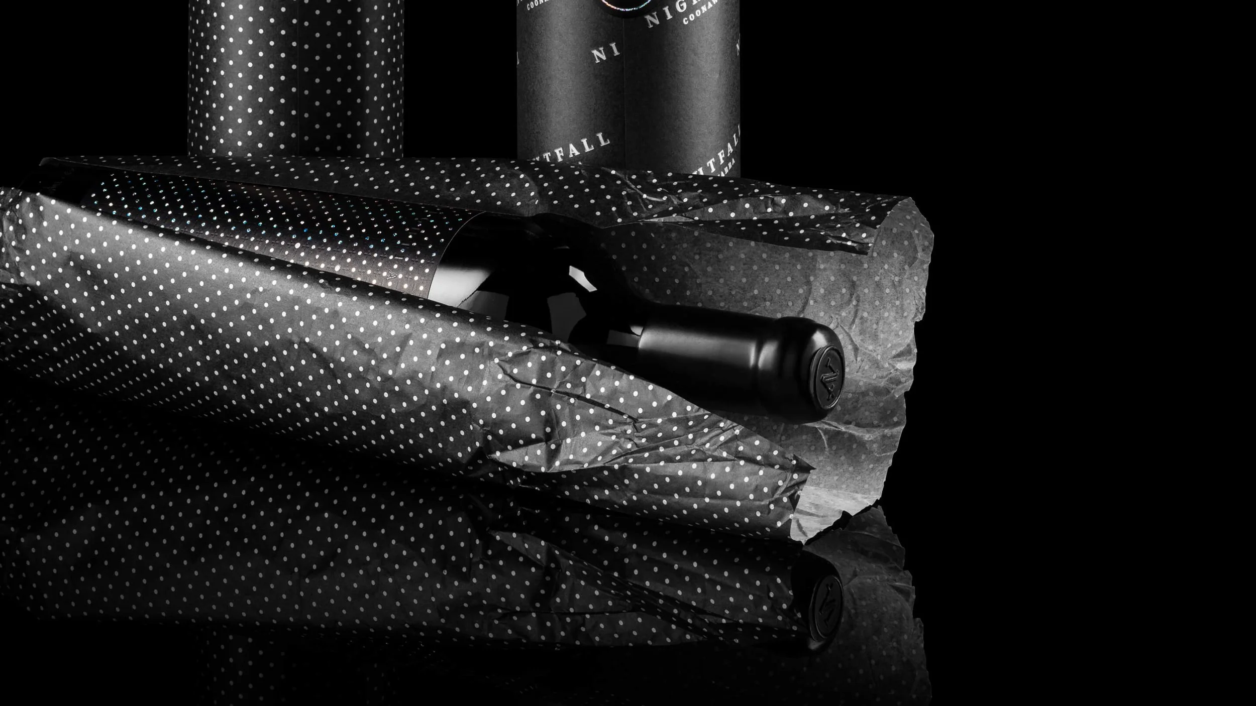 Black and white photograph of a wine bottle, a wrapped gift, and two cylindrical gift cans, all partially obscured by polka dot wrapping paper, against a dark background.