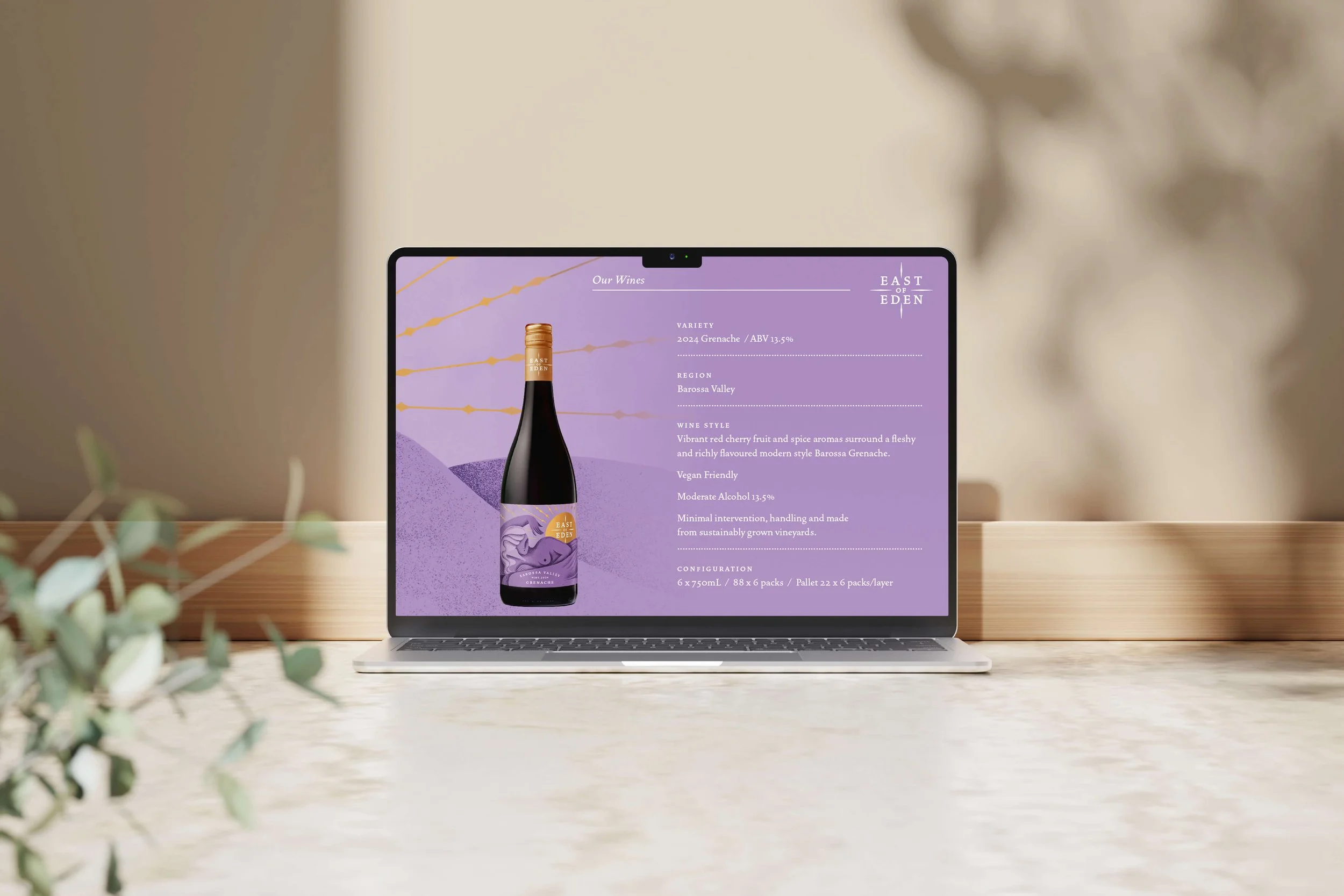 Laptop on a wooden table displaying a product page for a bottle of wine with a purple label and background. The wine is East of Eden, a Grenache variety from the Barossa Valley, with description of flavour notes, alcohol, and packaging details.