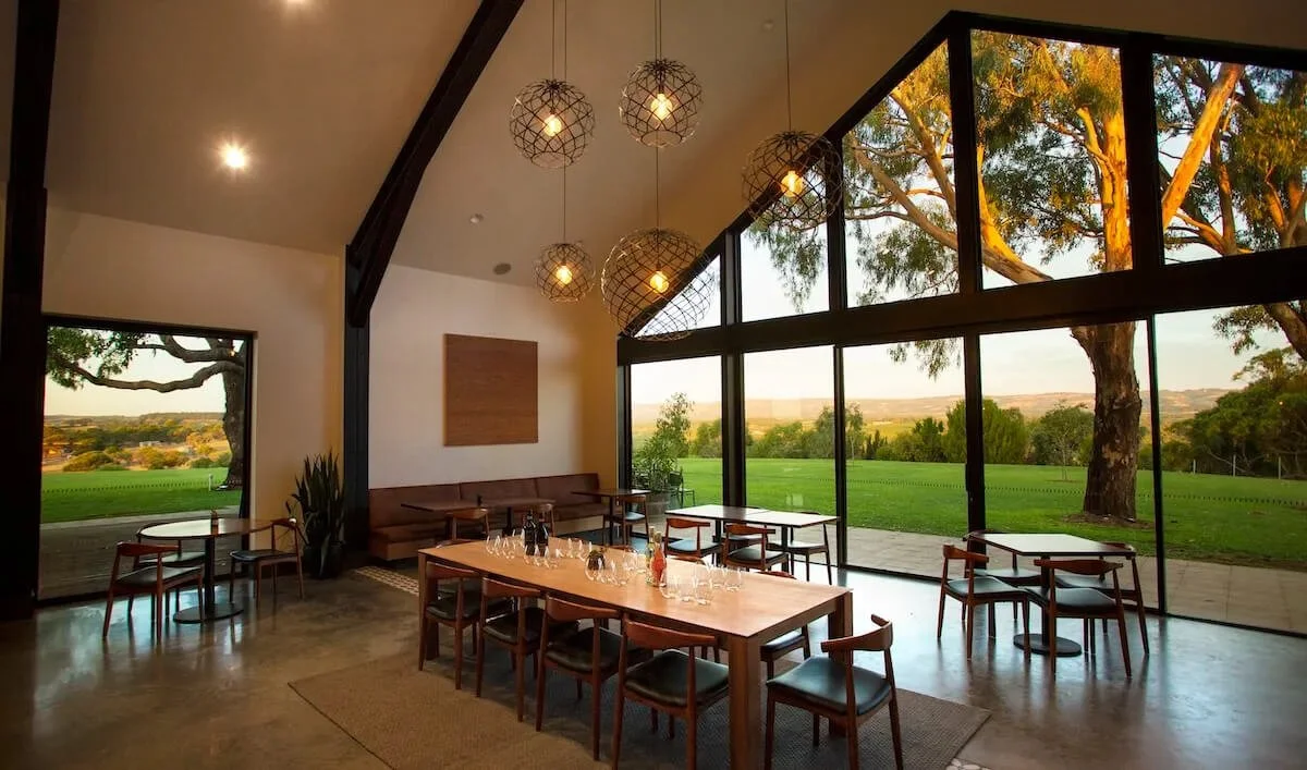 Internal image of Chalk Hill Wines modern dining room with large floor-to-ceiling windows overlooking a green outdoor landscape, with trees and rolling fields, featuring a wooden table set for a meal, hanging light fixtures, and minimalistic decor.