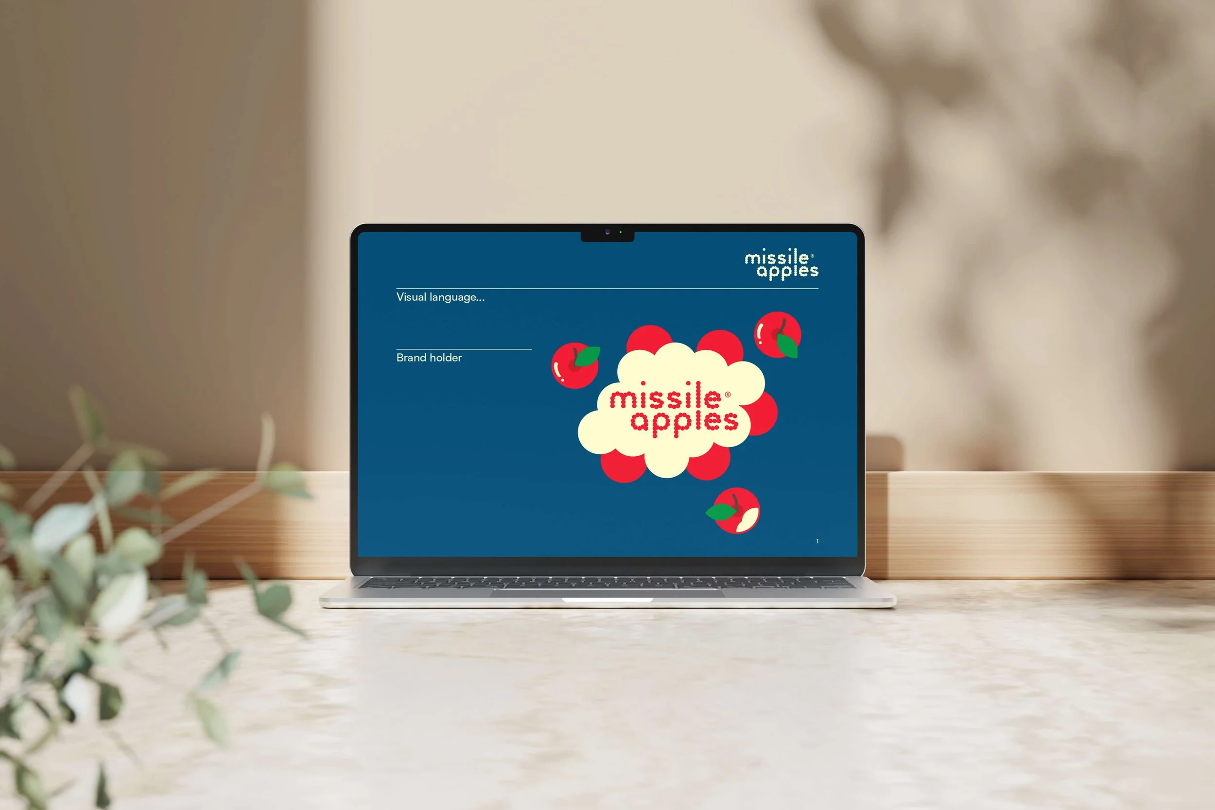 Laptop displaying a colorful graphic with red apples and a cloud with the words "missile apples" in red text, set on a wooden surface with a blurred leafy plant in the foreground.