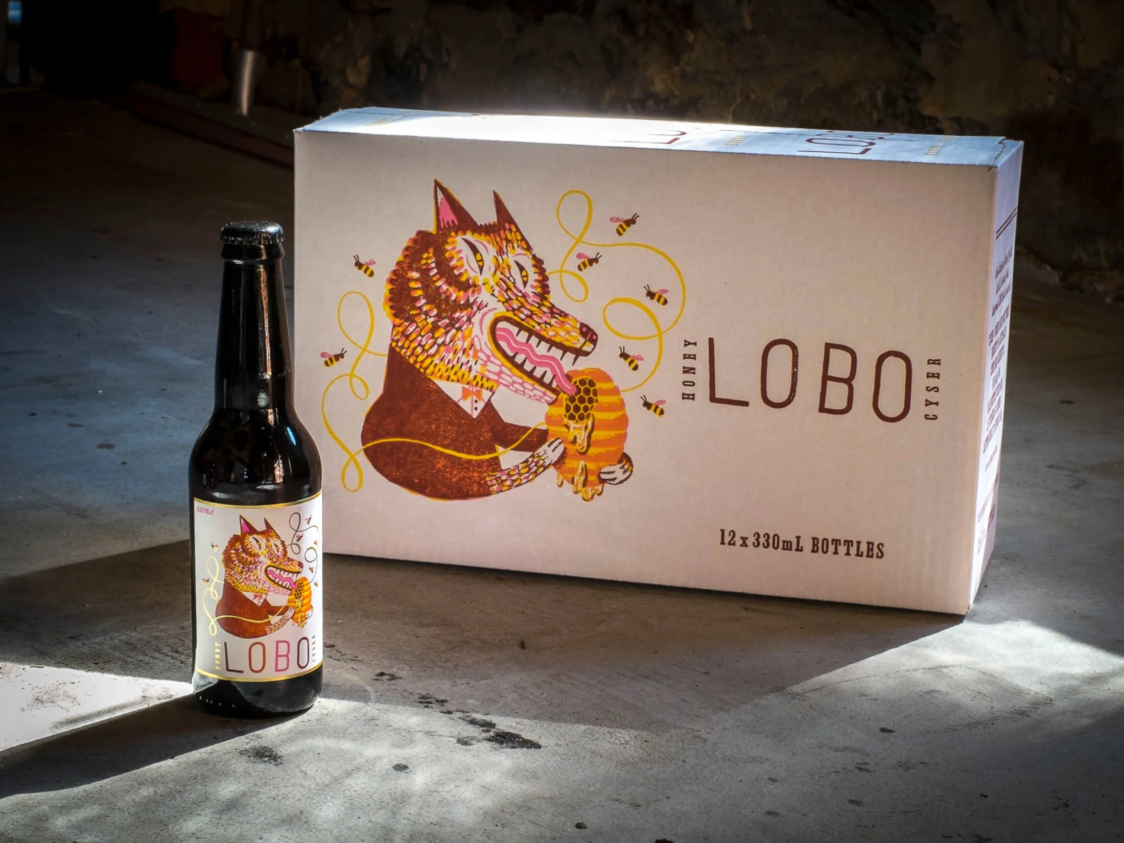 A six-pack of Lobo beer bottles with a box and a single beer bottle with a wolf illustration holding honey on a concrete surface.