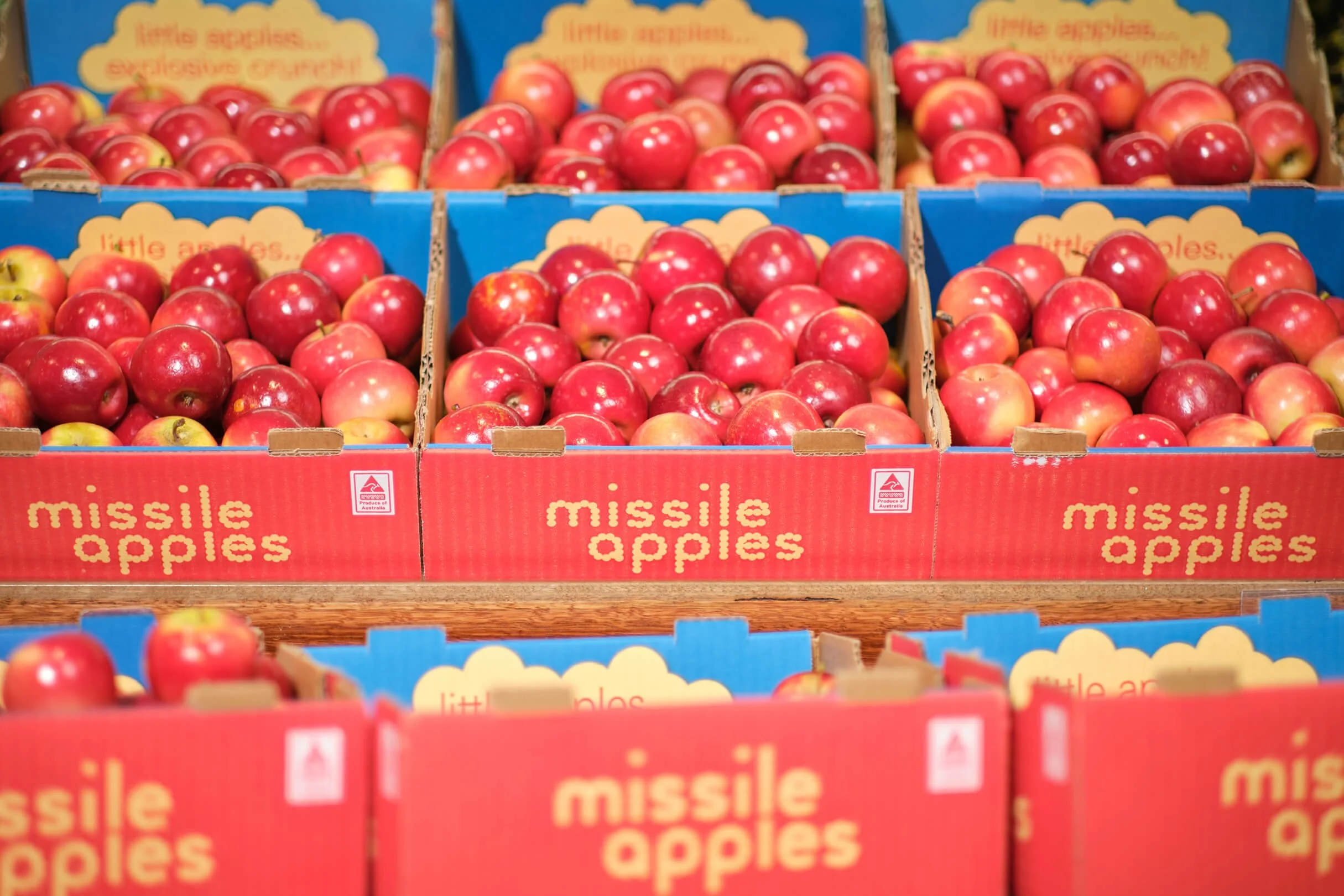 Multiple boxes of red and yellow apples labeled 'missile apples' arranged on a wooden surface at a market.