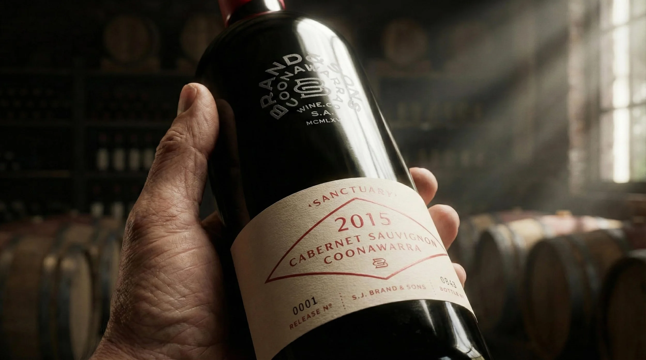 Person holding a bottle of red wine labeled 'Sanctuary 2015 Cabernet Sauvignon Coonawarra' inside a winery with wine barrels in the background.