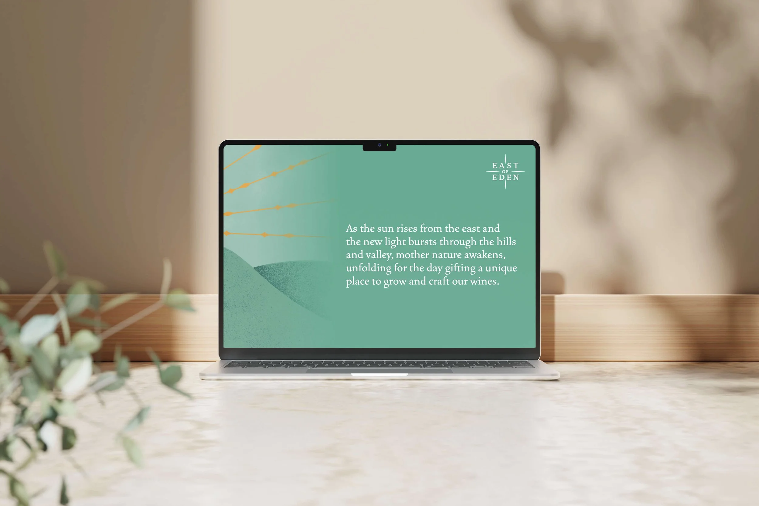 Laptop on a table displaying a green card with hills and sun rays, with text about the sunrise in the east and wine-making at East of Eden.
