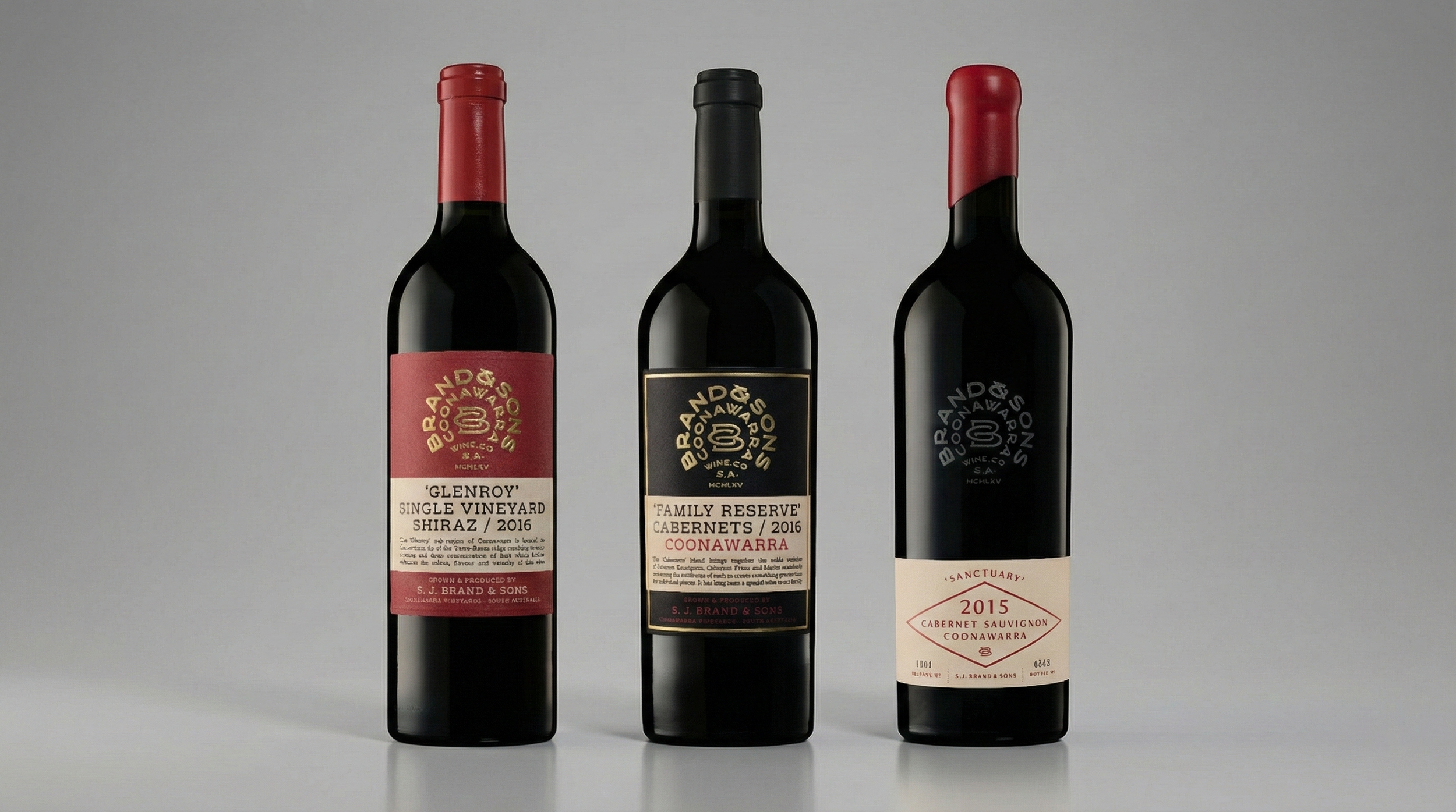 Three bottles of red wine standing upright against a plain background. The first bottle has a red label and cap, the second has a black label and cap, and the third has a white label and a red cap.