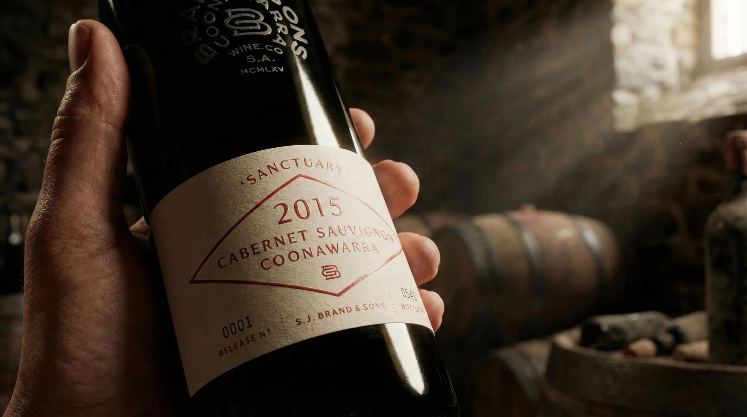Close-up of a hand holding a wine bottle labeled 'Sanctuary 2015 Cabernet Sauvignon Coonawarra' in a cellar with wooden barrels in the background.