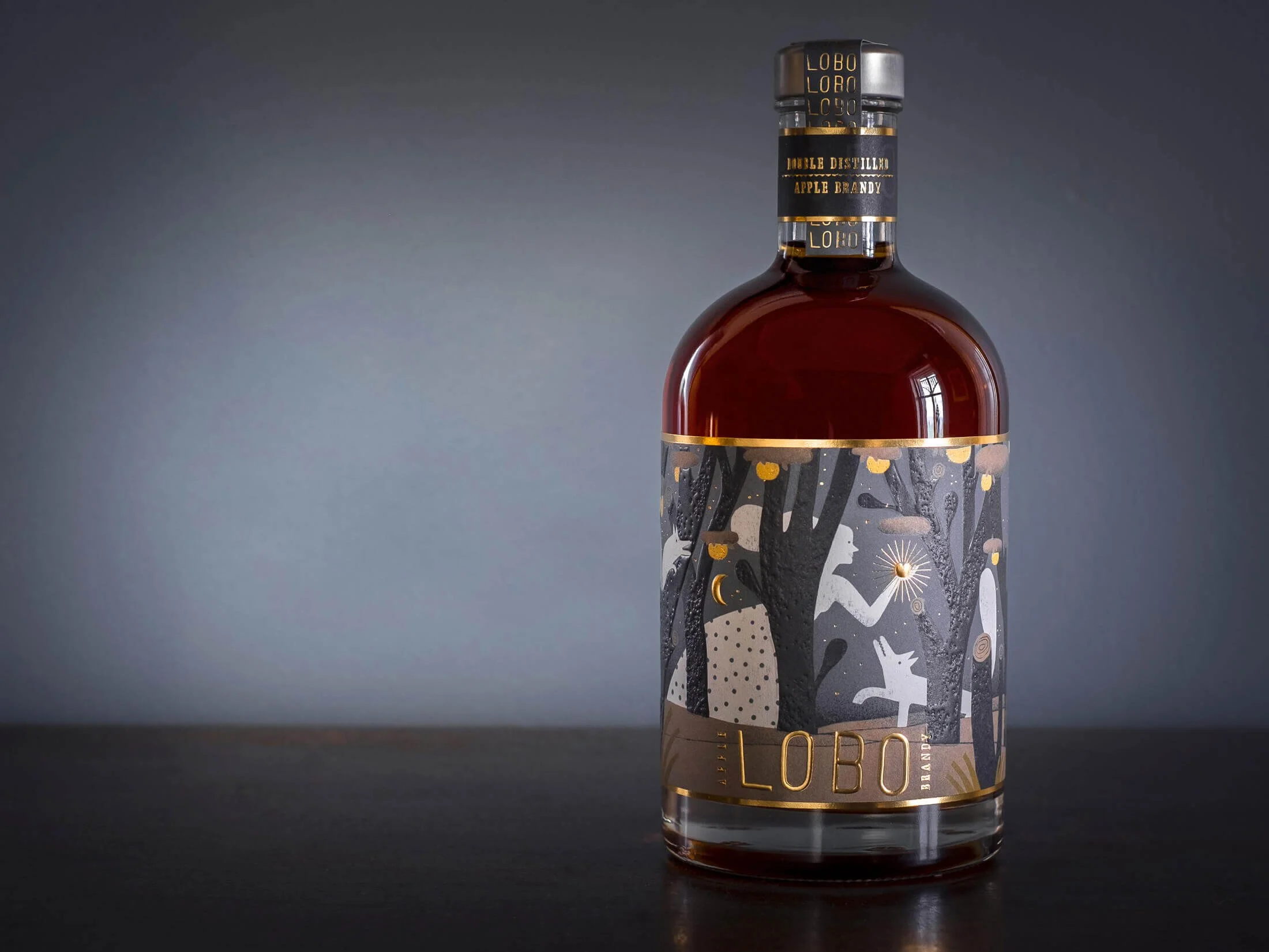 A bottle of Lobo brandy with a decorative label featuring black and gray trees, a white rabbit, and gold accents, set against a dark background.