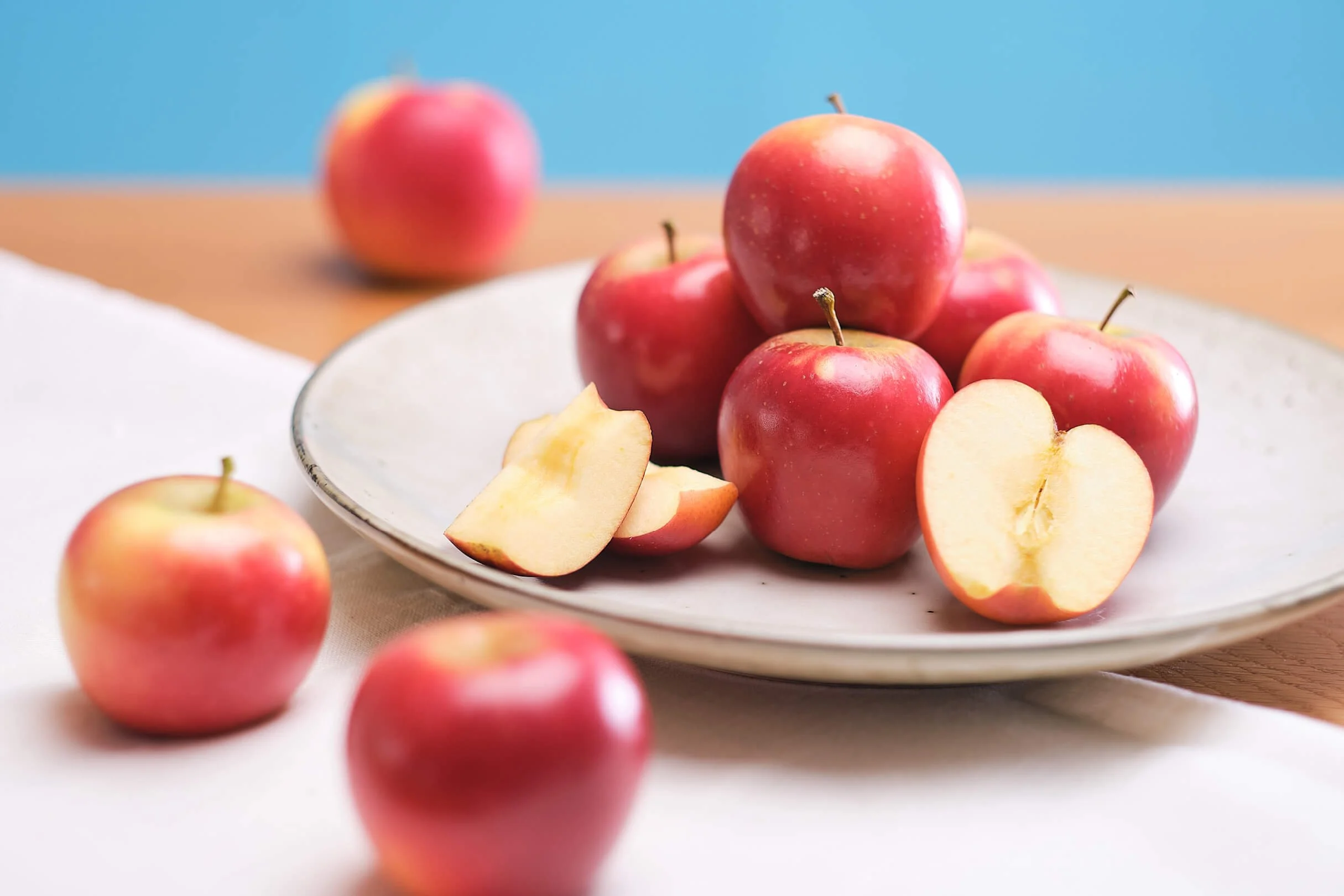 A white plate with red and yellow apples, some sliced, on a wooden table with a light blue background.
