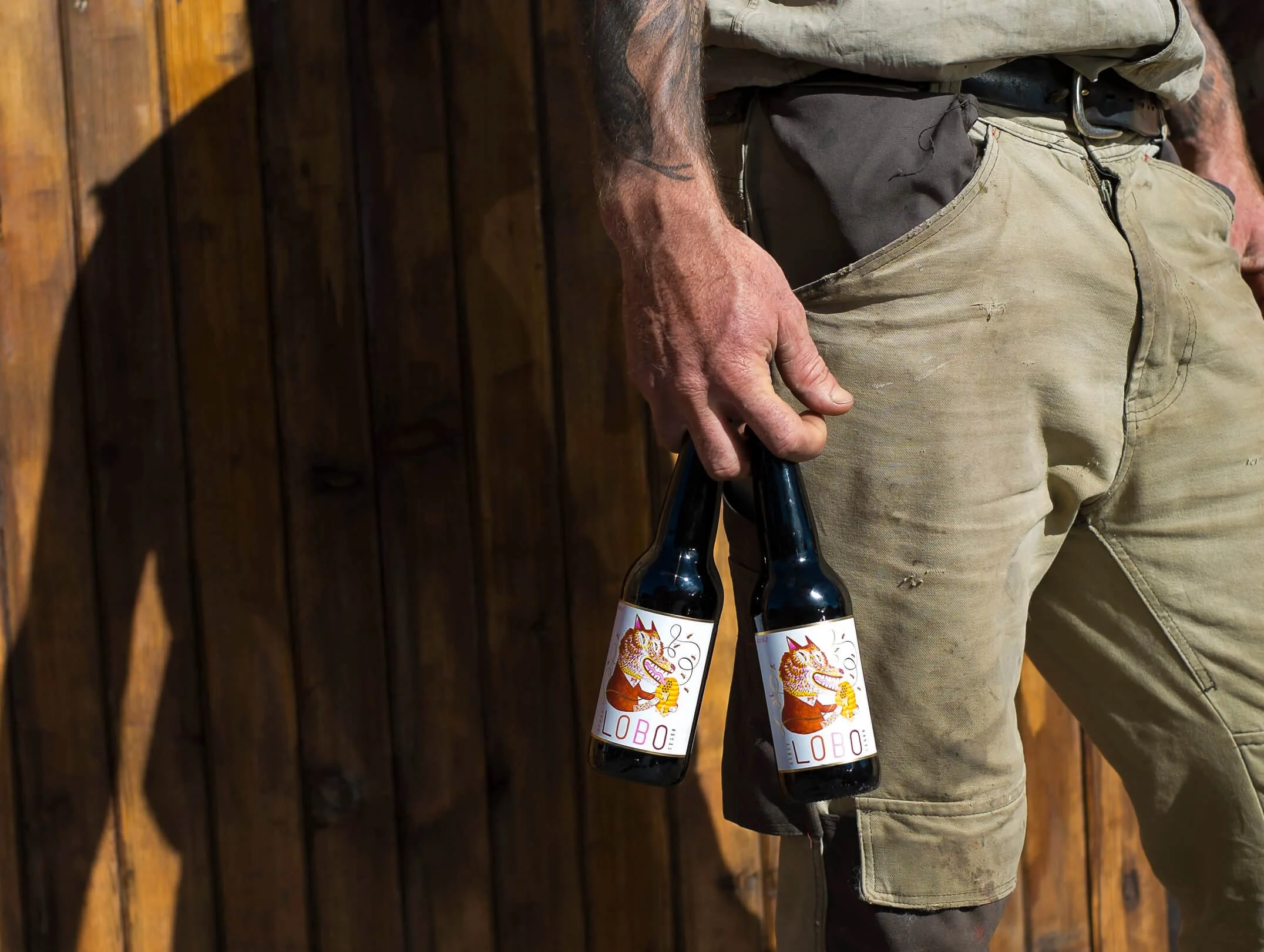 Person holding two beer bottles with a cartoon cat on the labels, standing in front of a wooden wall.