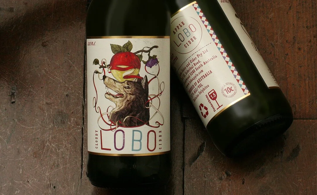 Two bottles of Lobo cider on a wooden surface, featuring artistic label designs with a wolf and a red apple.