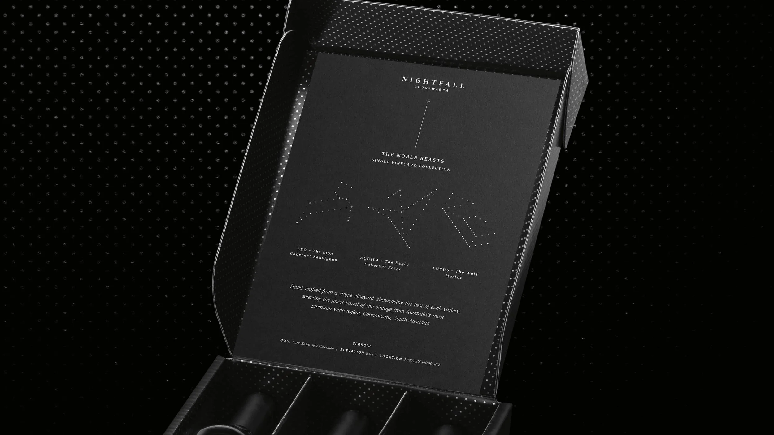 A black wine box with a transparent lid displaying three wine bottles inside. The box has a black interior with small white dot patterns. The label on the inside lid features a black background with white text and a star chart relating to wine varieties.