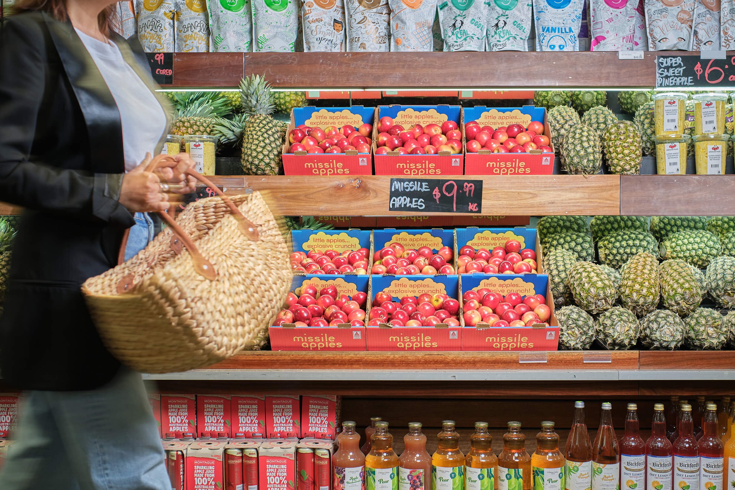 A shopper with a woven basket shopping for fruit at a grocery store. The produce section features trays of red apples labeled 'missile apples' and pineapples on the side. There are beverage bottles on the lower shelf.