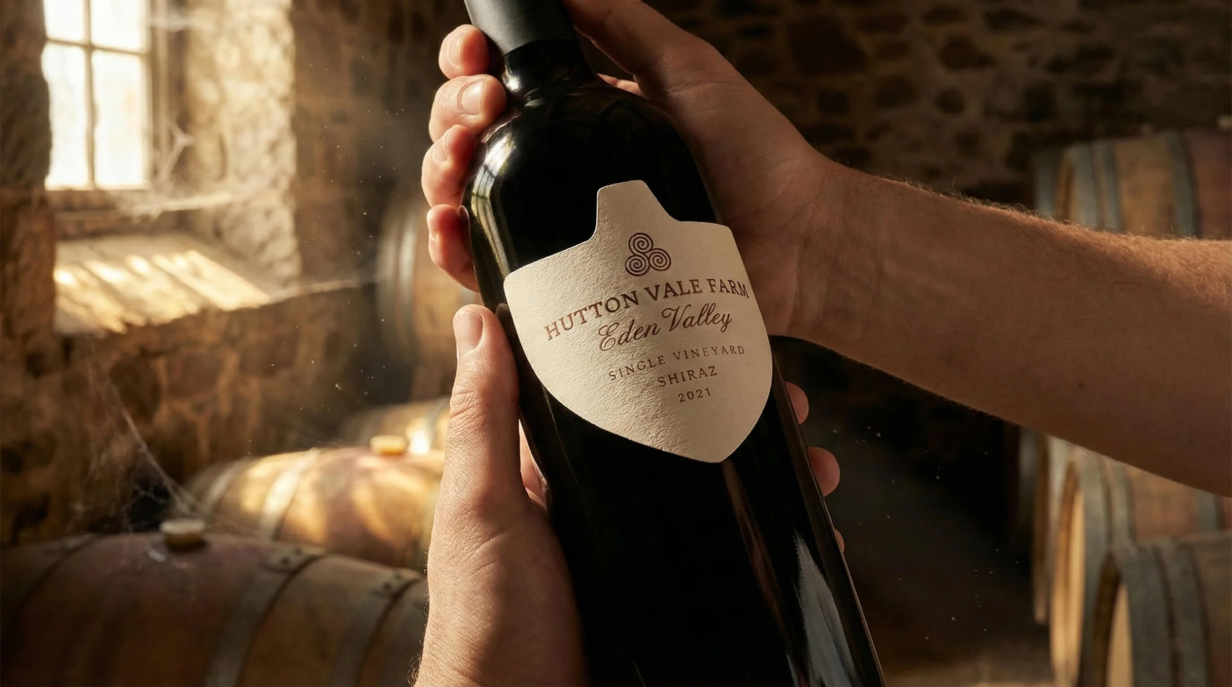 Person holding a bottle of Hutton Vale Farm Eden Valley single vineyard Shiraz 2021 wine inside a rustic wine cellar with barrels and a window in the background.