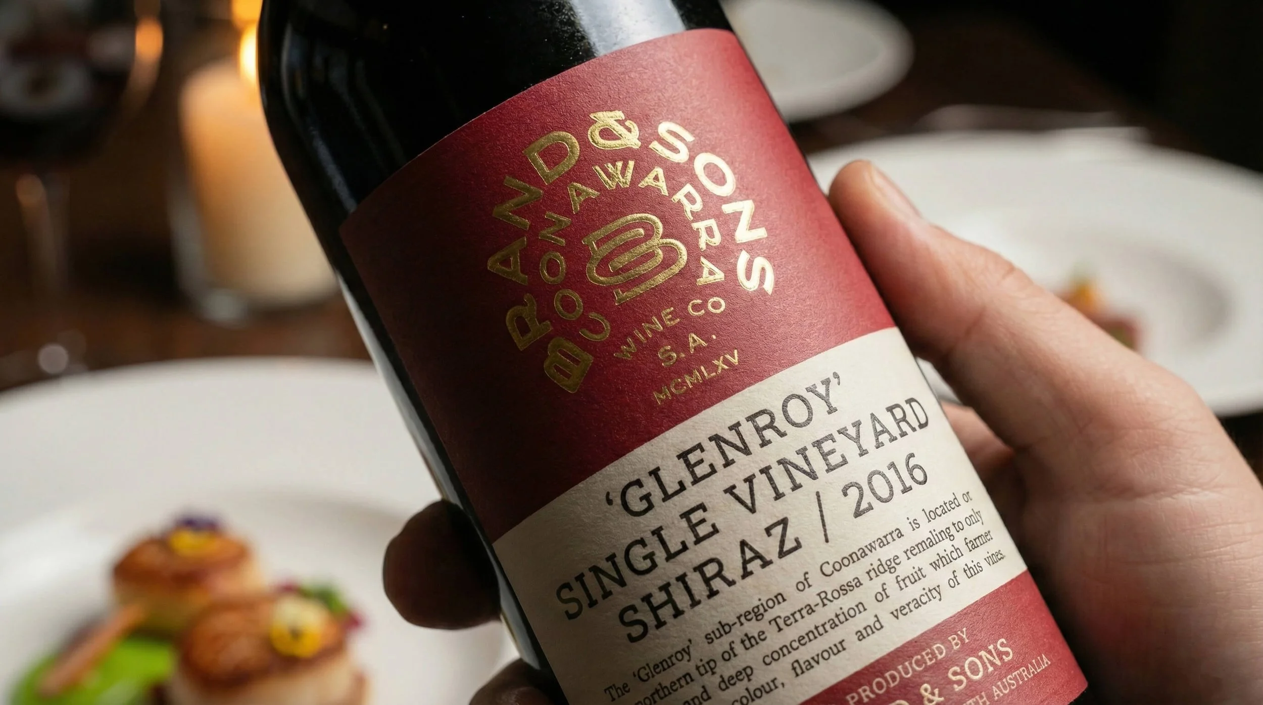 A hand holding a bottle of Windpound Wines' Glenroy Shiraz 2016, with a blurred background of a restaurant table and plates of food.
