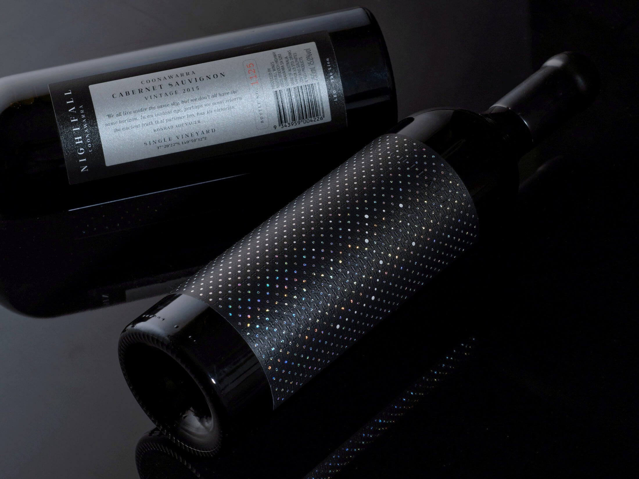Nightfall Wines claret shaped wine bottle laying on its side with subtle light falling on to it. Shows a black front label with star constellations. Back silver label with wine information.