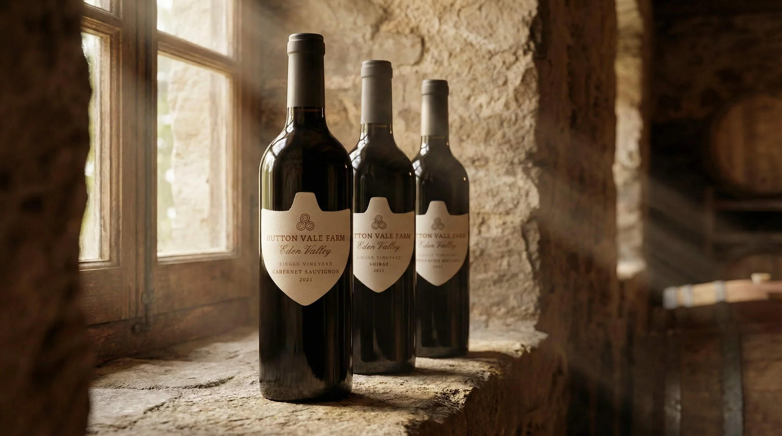 Three bottles of red wine from Hutton Vale Farm, Eden Valley, single vineyard, Cabernet Sauvignon and Shiraz, vintage 2021, are placed on a rough stone windowsill in a rustic room with exposed stone walls and wooden window frames.