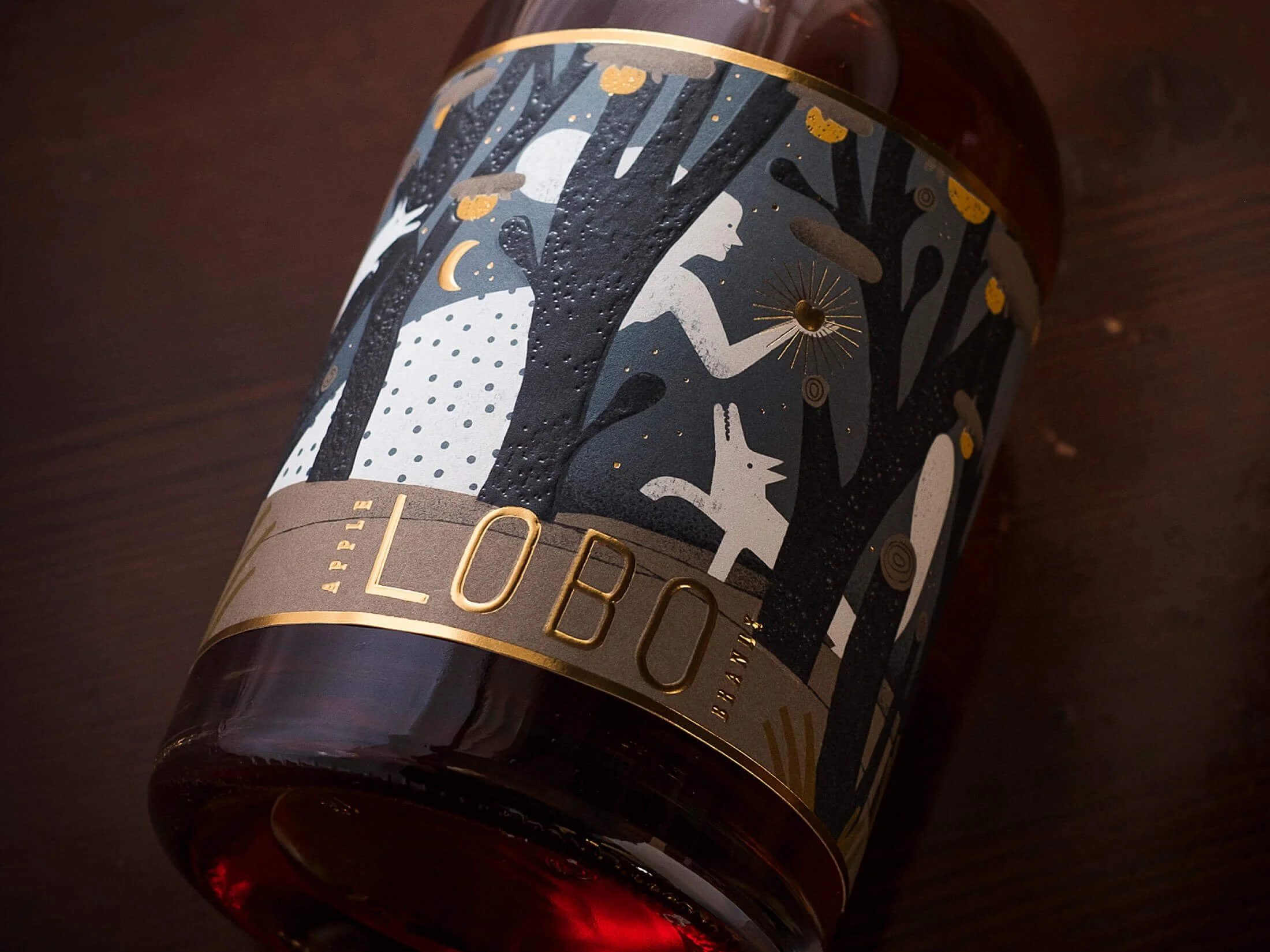 A dark glass bottle with a label depicting an illustrated forest scene with trees, a woman, a rabbit, and animals, featuring gold accents and the brand name 'Lobo' in gold lettering.