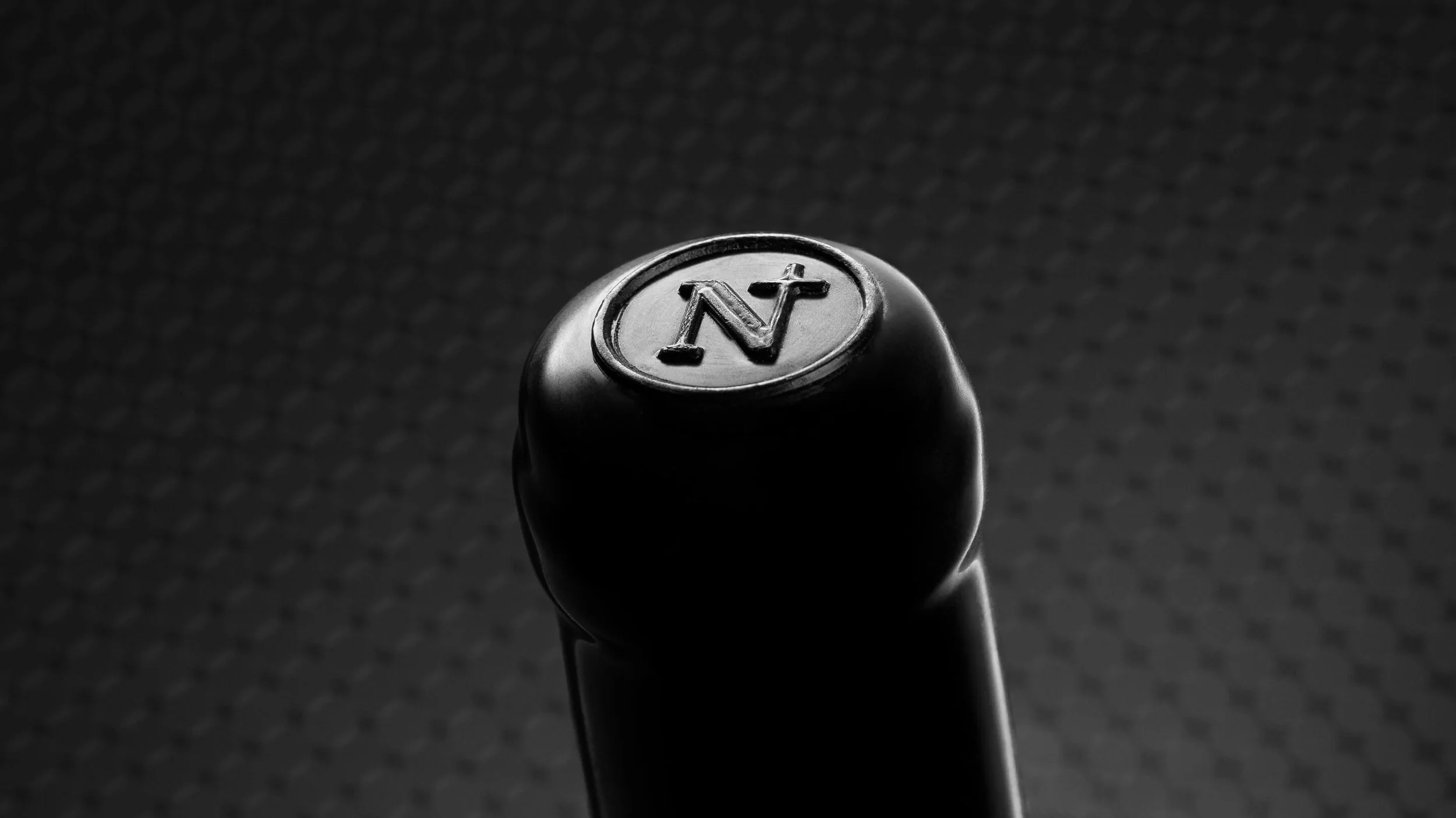 Close-up of a black wax capped wine bottle with a circular top imprinted with the letter 'N' inside an oval, against a dark textured background for Nightfall Wines.