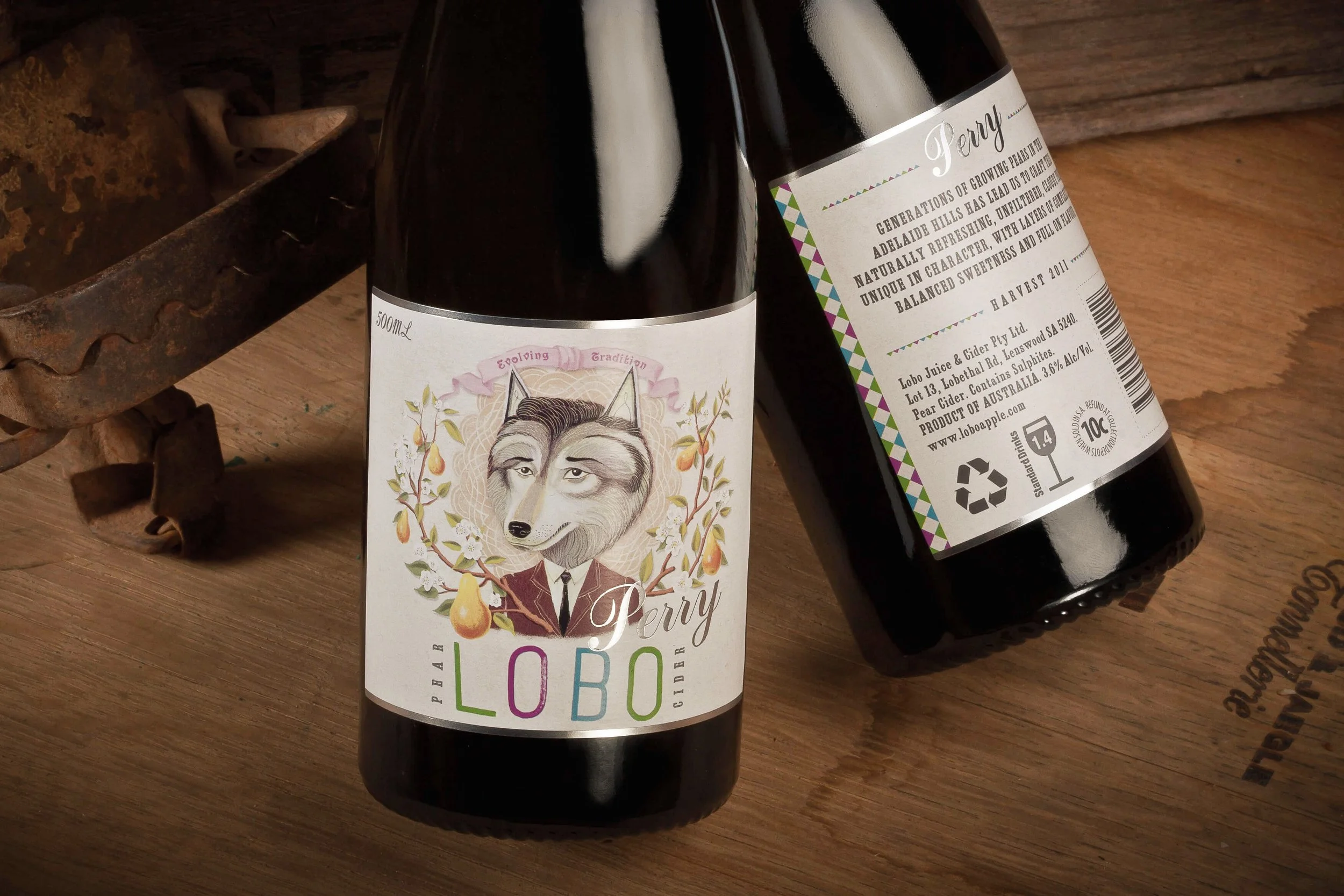Two dark glass bottles of Lobo Perry cider on a wooden surface, one showing front label with a wolf illustration and colorful text, the other showing back label with product details