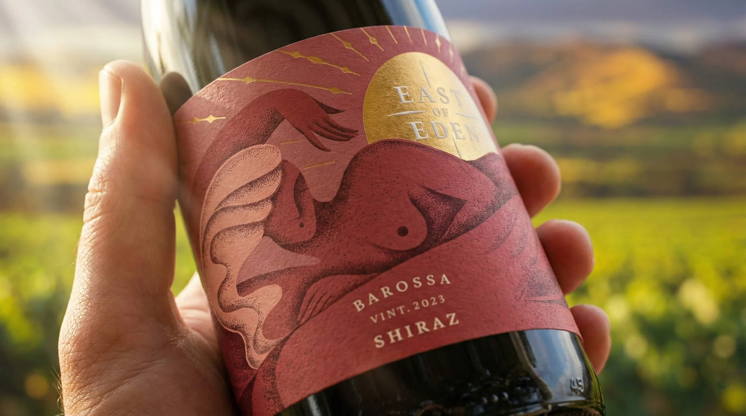 Close-up of a hand holding a wine bottle with a pink and gold label featuring an abstract illustration of a woman and a yellow sun, reading 'East of Eden,' 'Barossa,' 'Vint. 2023,' and 'Shiraz,' with a blurred vineyard landscape in the background.