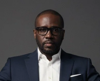 A man with glasses dressed in a dark suit and white shirt against a plain gray background.
