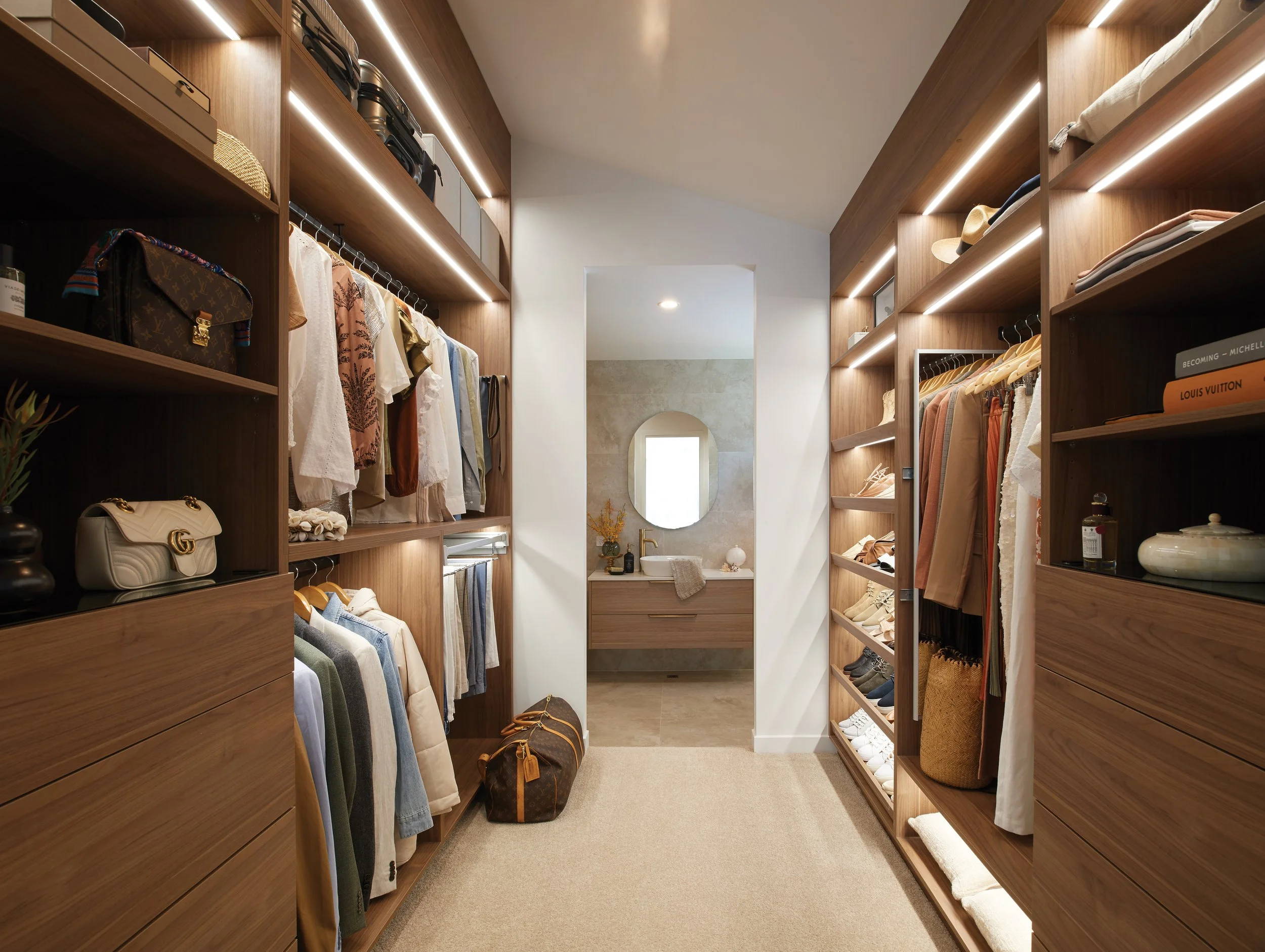 Walk-in closet with wooden shelves and clothing, bags, shoes, and accessories, leading to a bathroom area with a vanity, mirror, and natural light.
