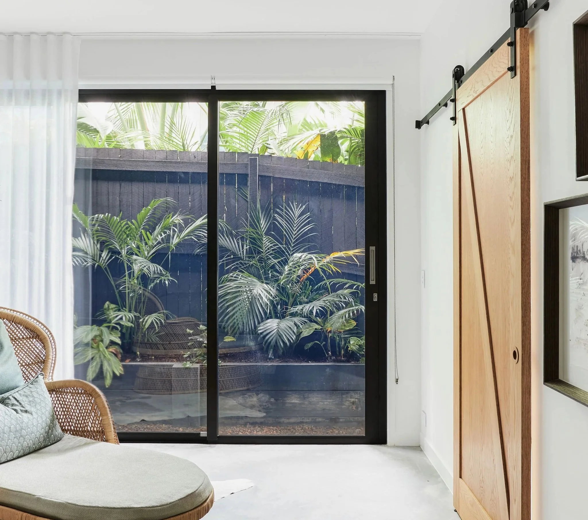 Interior room with white walls, sliding glass door revealing plants outside, rattan chair with cushions, wooden sliding barn door on right wall, and framed artwork on the wall.