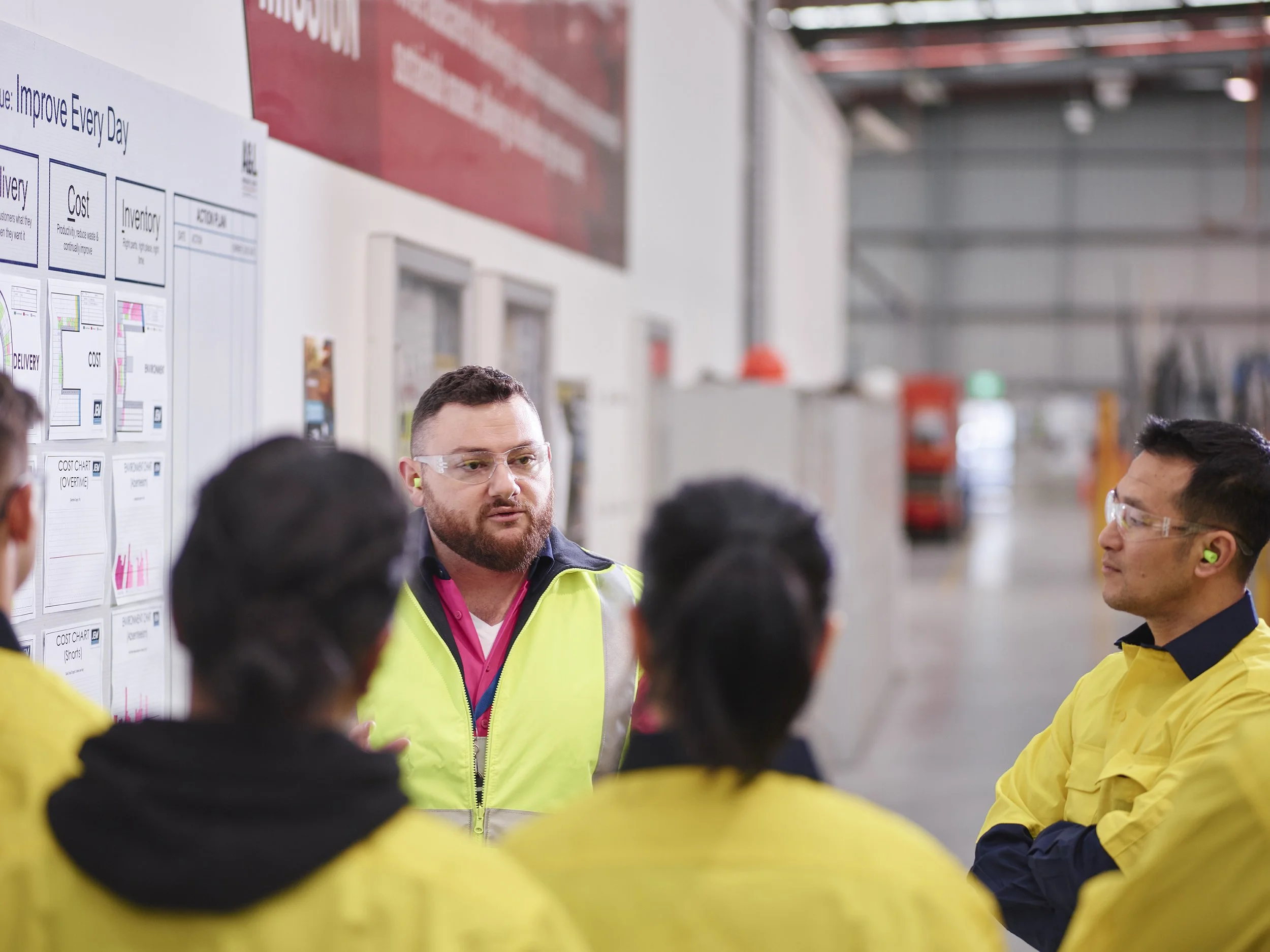 A group of workers in yellow safety gear listening to a man in a high-visibility vest giving a safety briefing in an industrial warehouse.