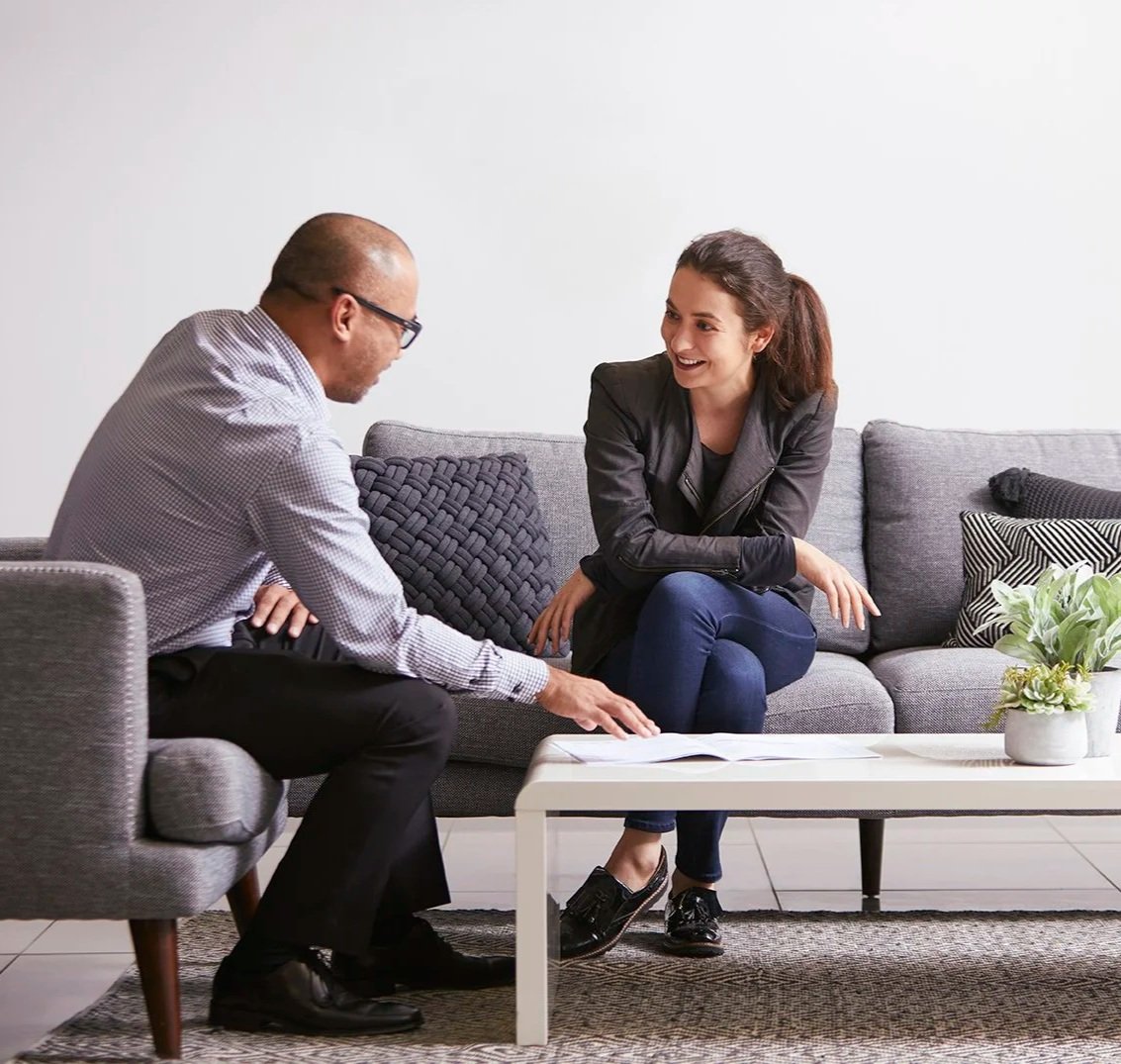 A man and woman sitting on a gray sofa, engaged in conversation, with the woman smiling, in a modern living room.