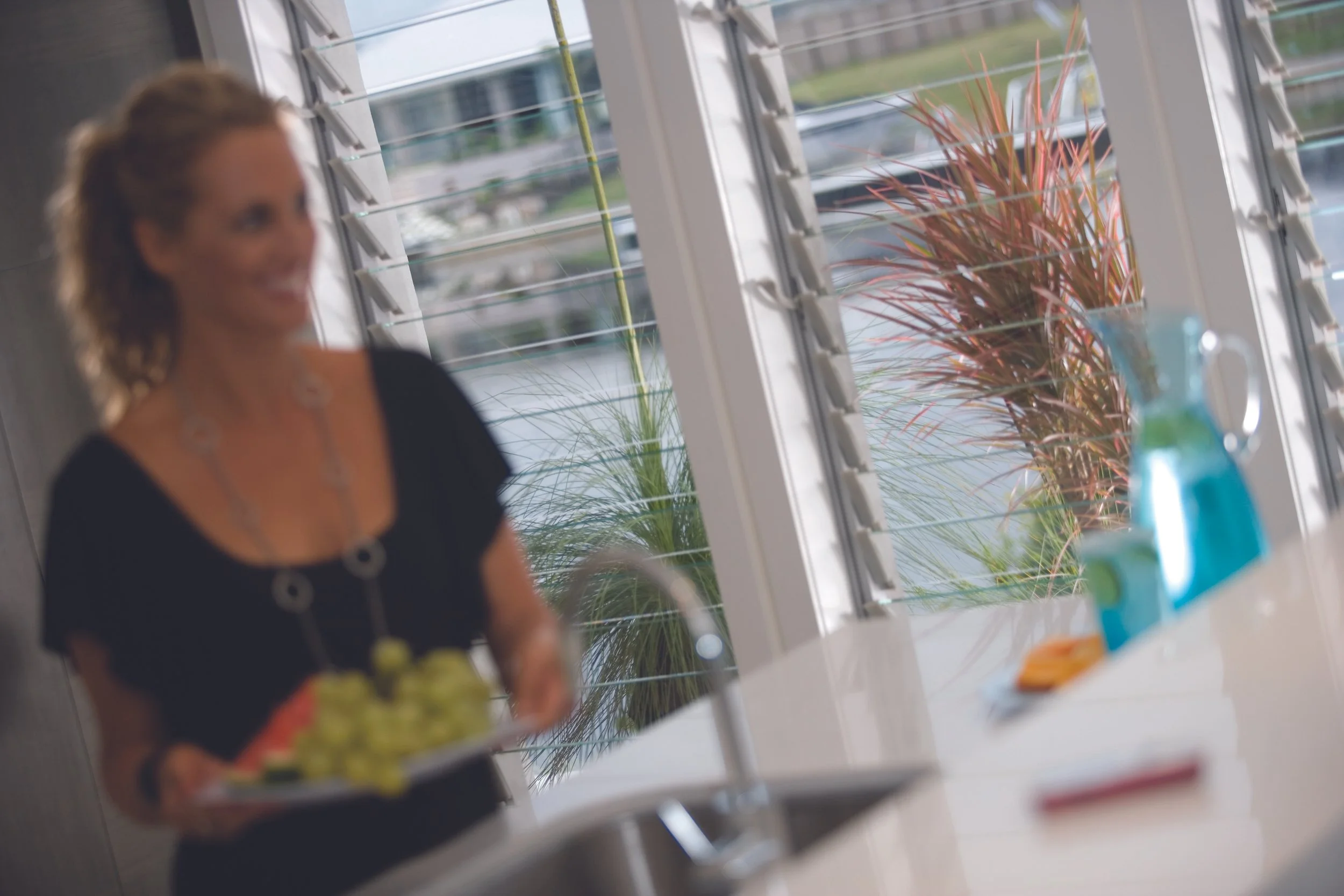 A woman in a black top holding a plate of food standing in a bright kitchen near large windows with outdoor plants.