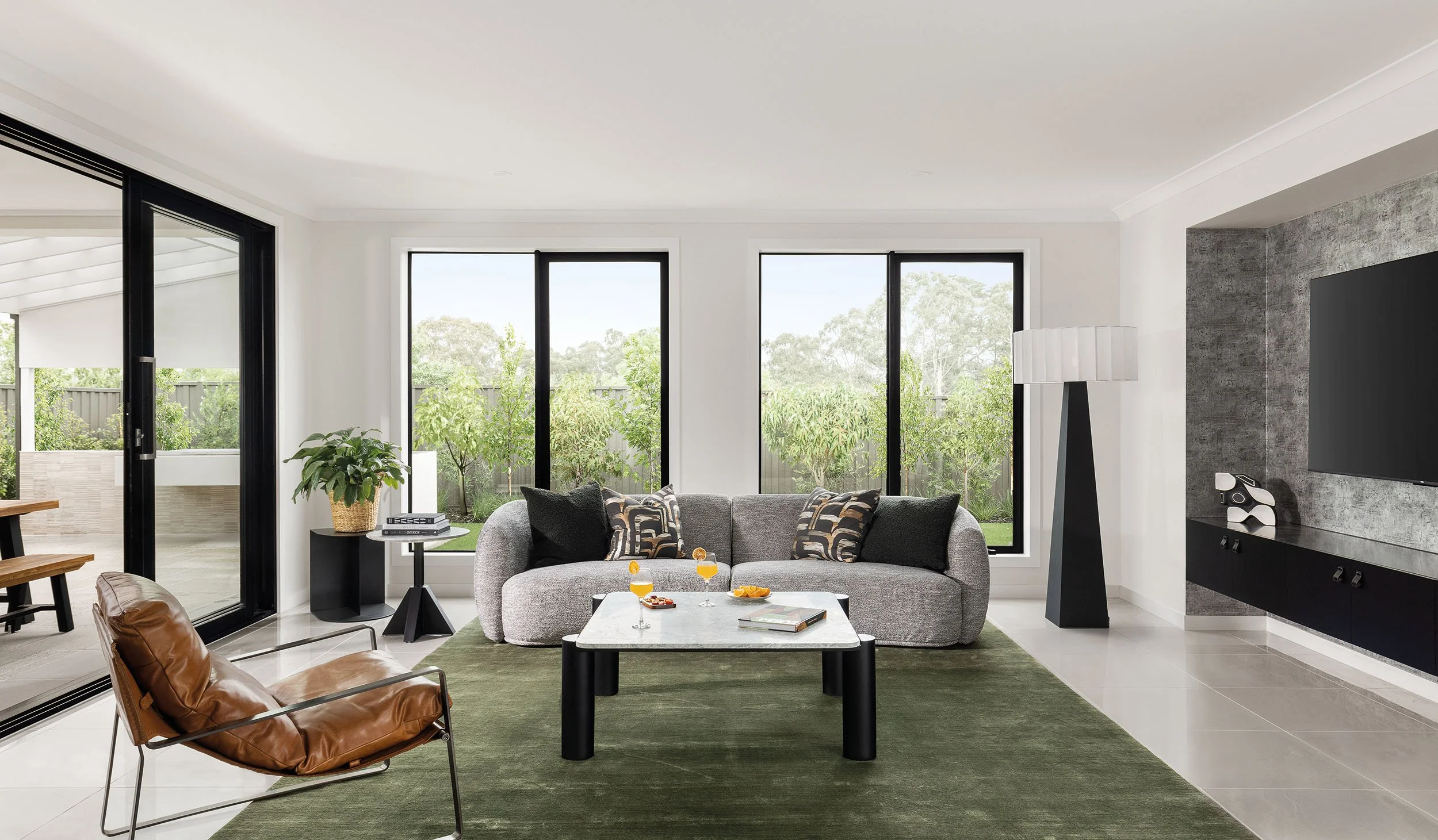 Modern living room with gray sofa, glass coffee table, leather lounge chair, large windows overlooking garden, and TV mounted on a textured wall.