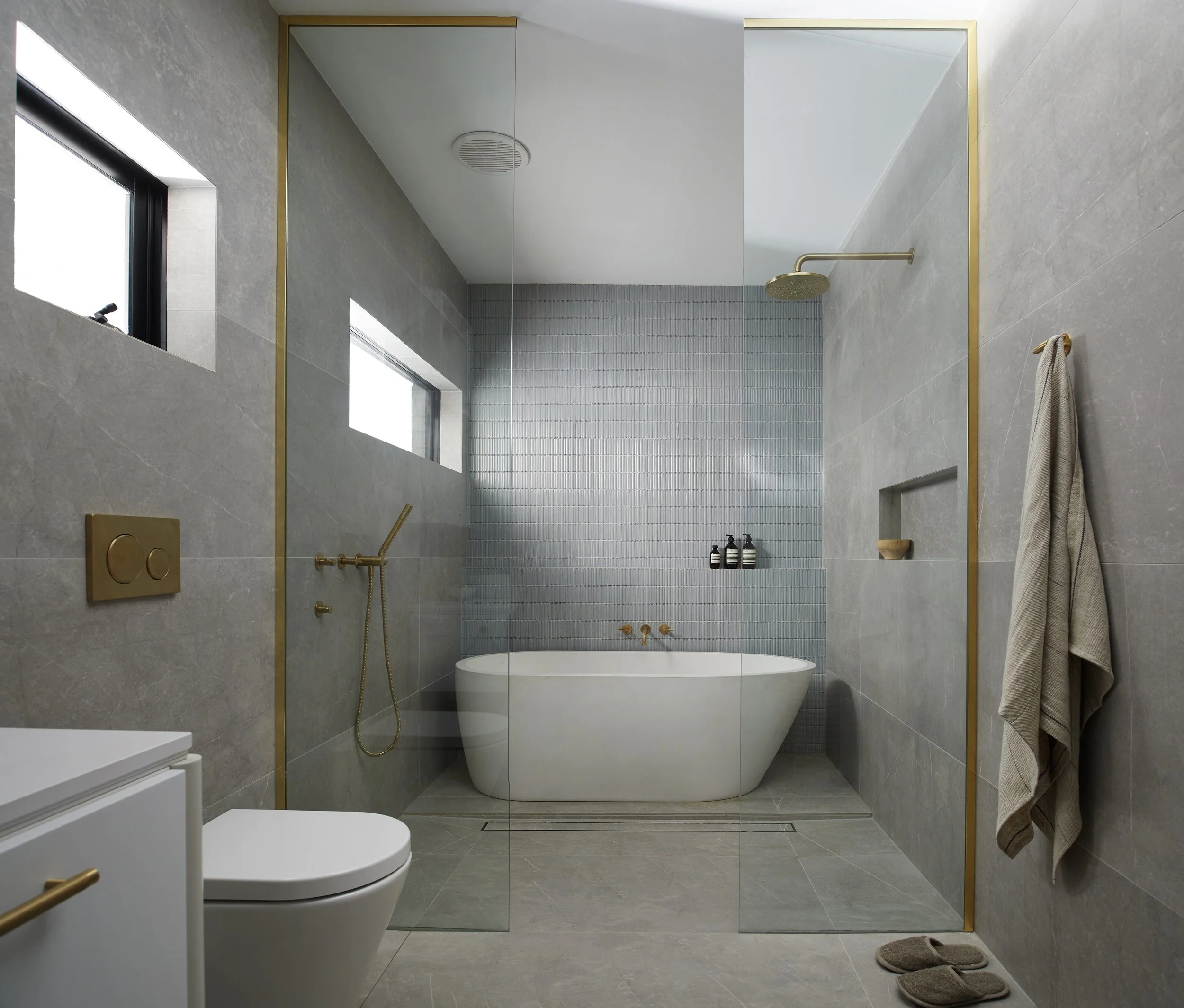 Modern bathroom featuring a bathtub, gray tiled walls, a glass shower partition with gold trim, and minimalistic fixtures.