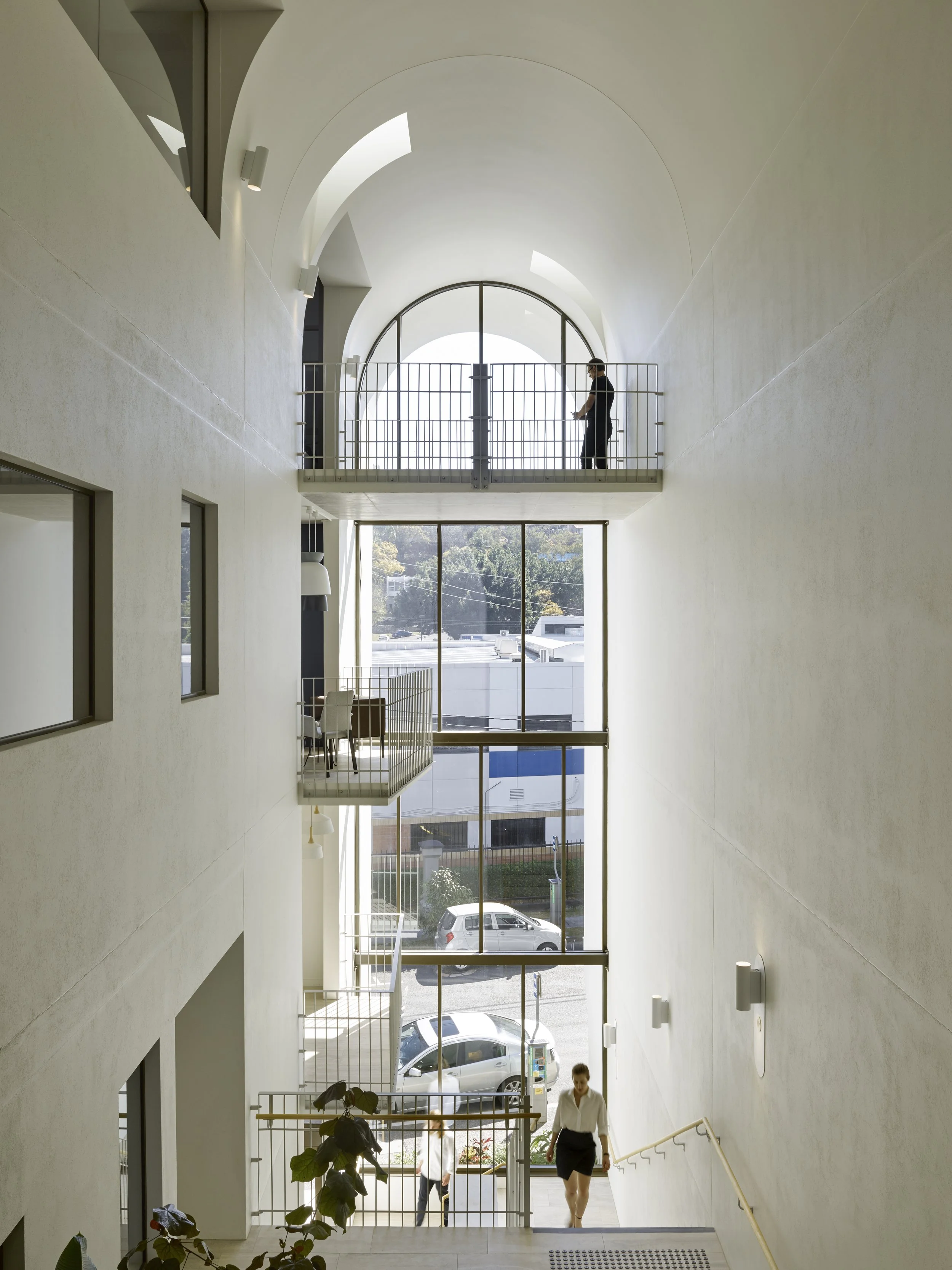 Interior of a modern building with multiple floors, large windows, and two people walking, one on each upper and lower levels.